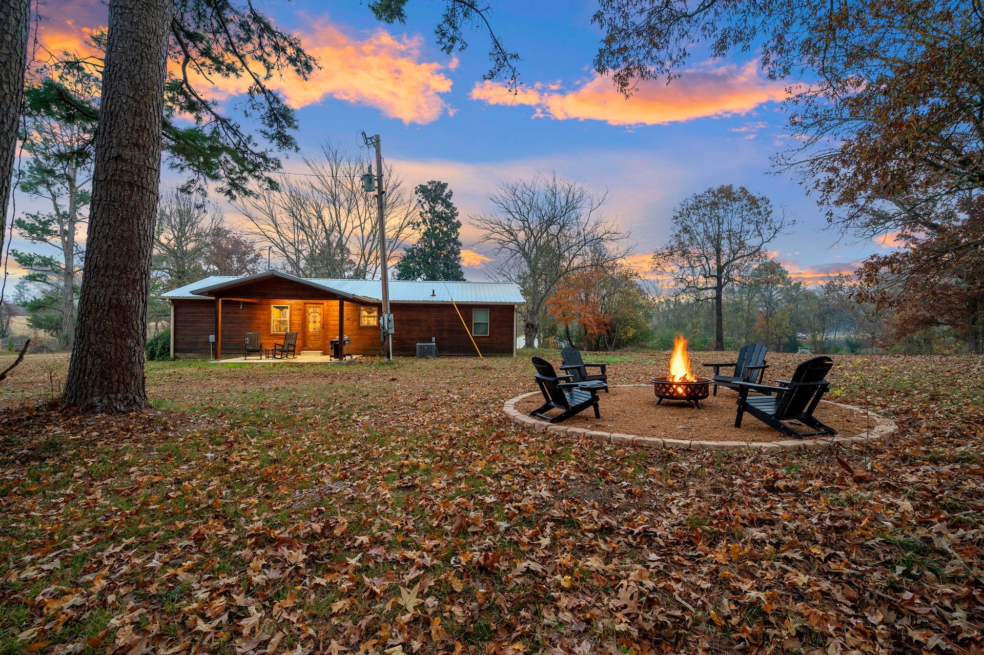A cabin sits on a leaf-covered lawn at twilight, with a fire pit surrounded by chairs in the foreground.