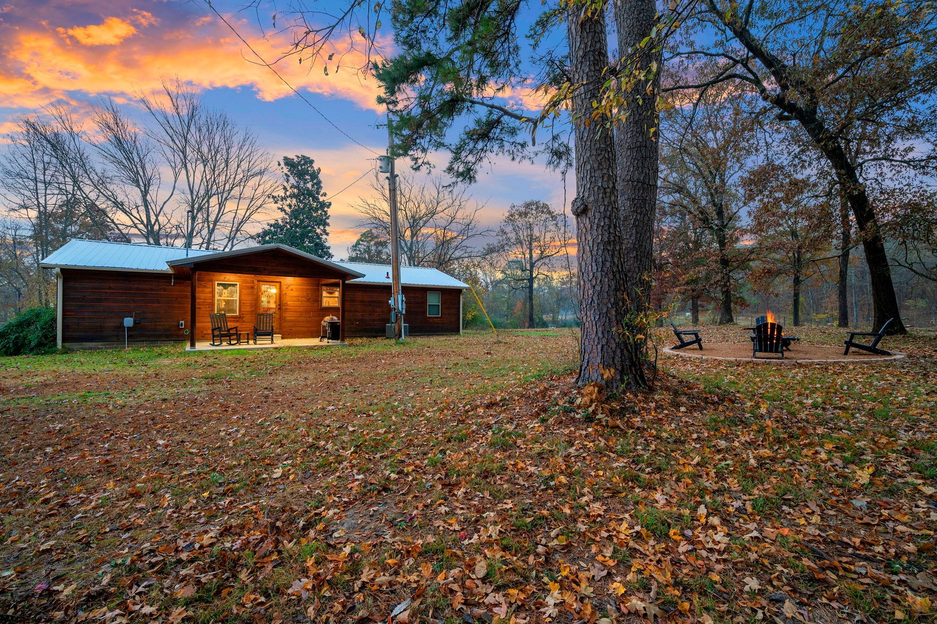 A wooden cabin sits in a clearing covered in autumn leaves, with a fire pit nearby under a colorful sunset sky.