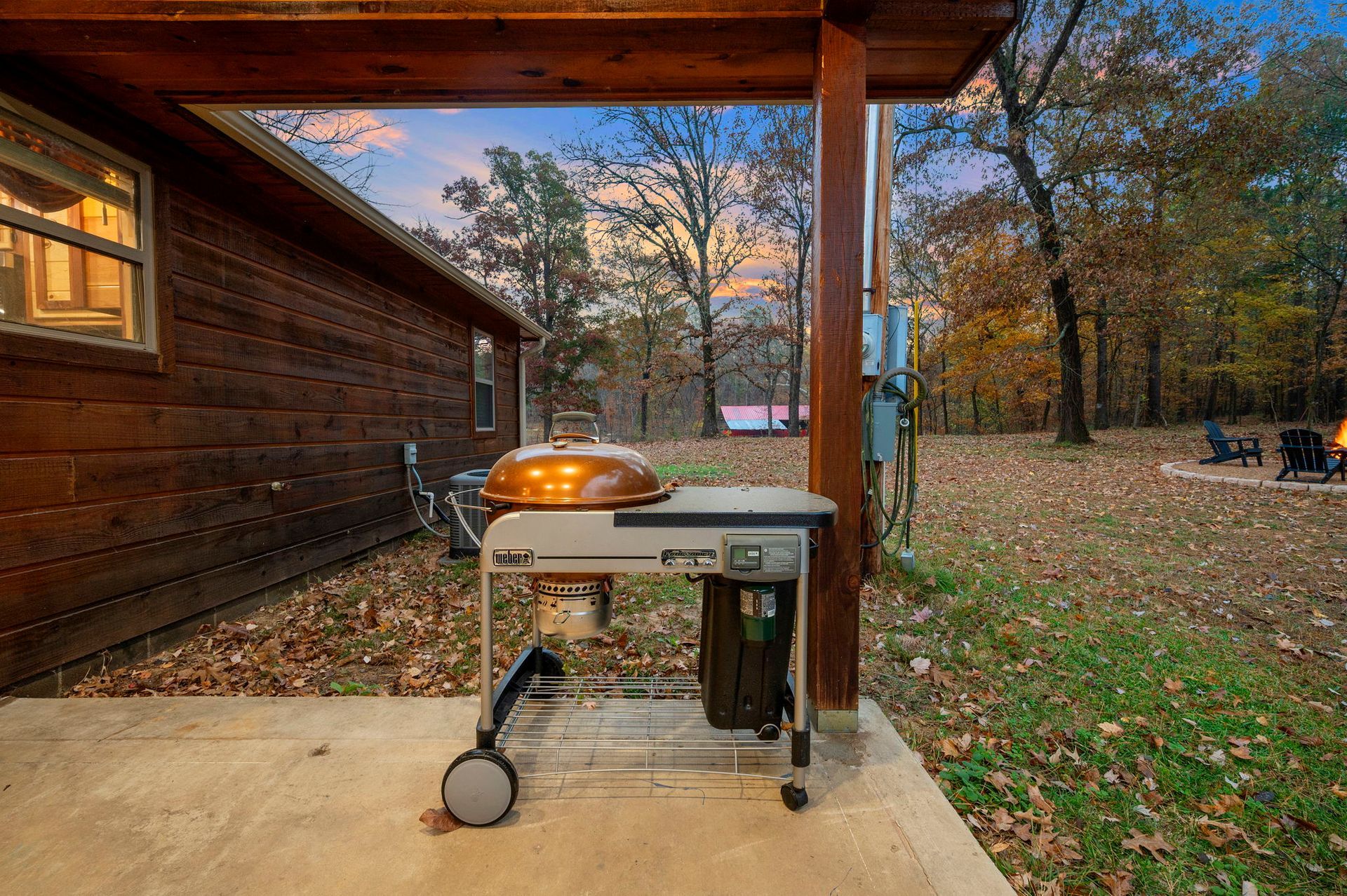 A copper-lidded grill sits on a concrete patio under a wooden cabin awning, overlooking a wooded yard at dusk.
