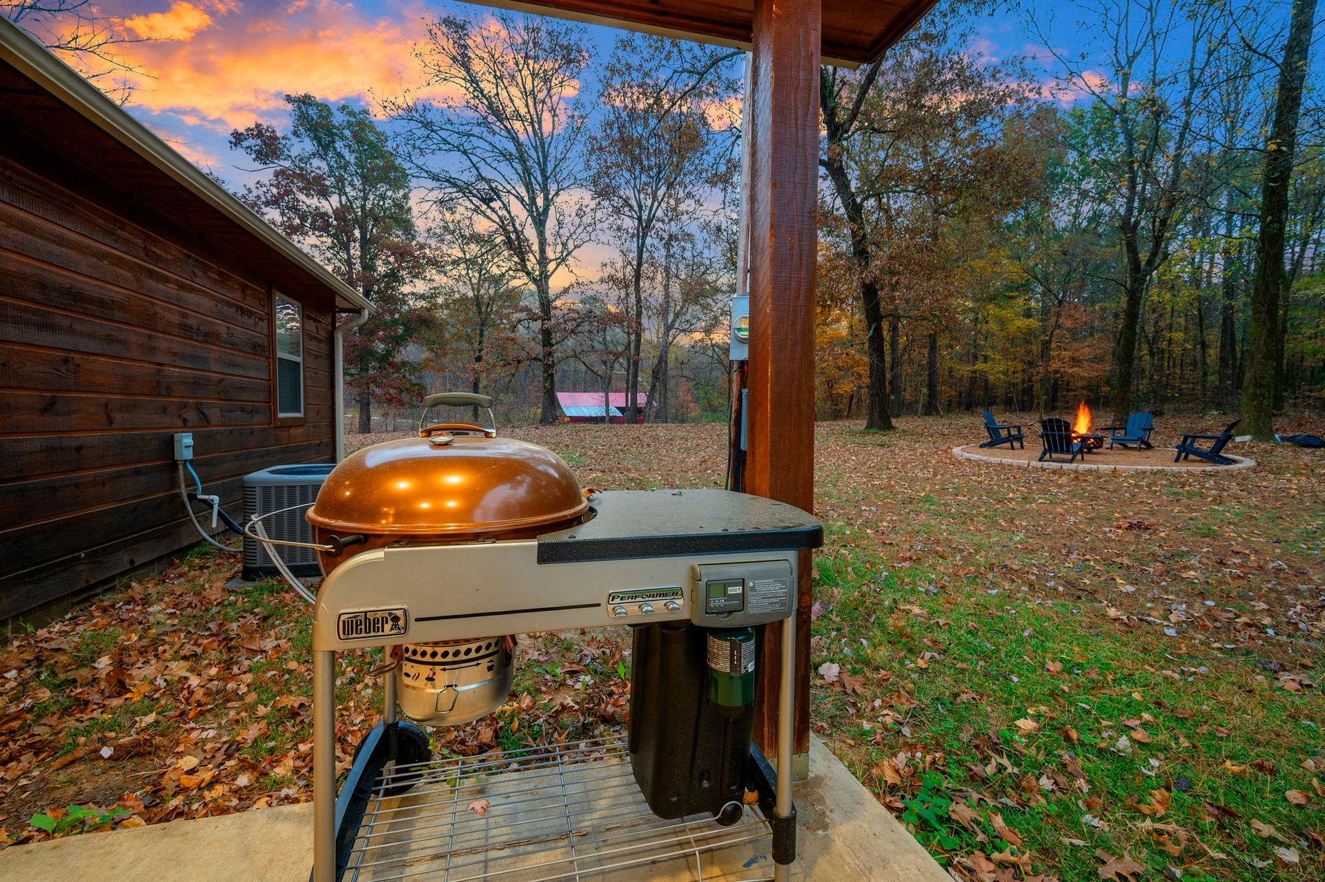 A bronze grill sits on a patio next to a wooden cabin, overlooking a backyard fire pit at sunset.