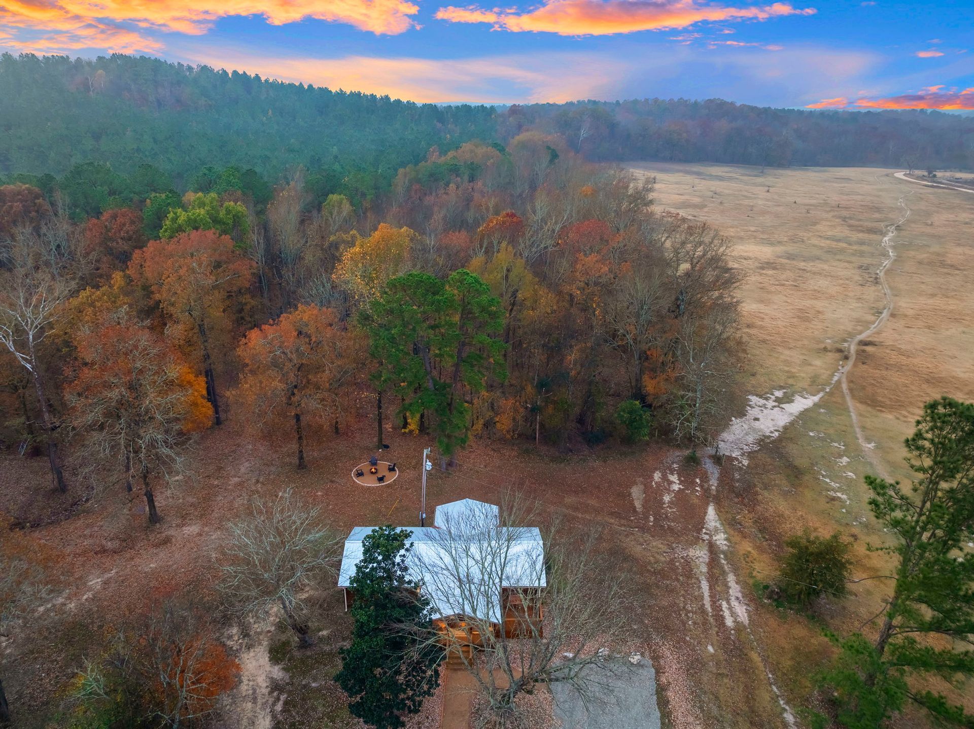 Aerial view of a cabin with a metal roof nestled beside a forest and field at sunset.