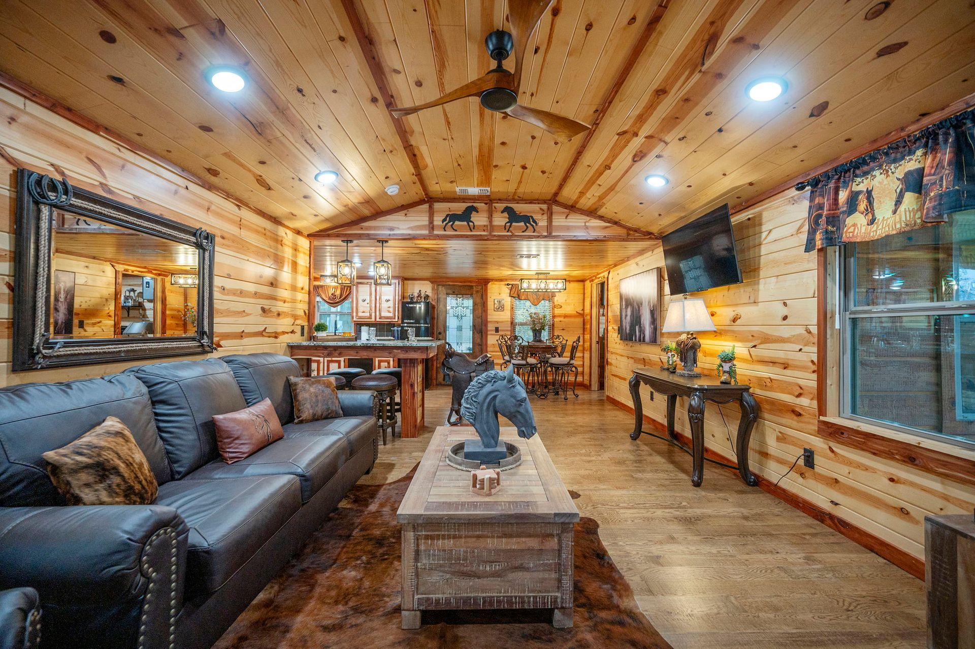 A rustic living room with pine walls, a black leather sofa, a wooden coffee table, and a ceiling fan.