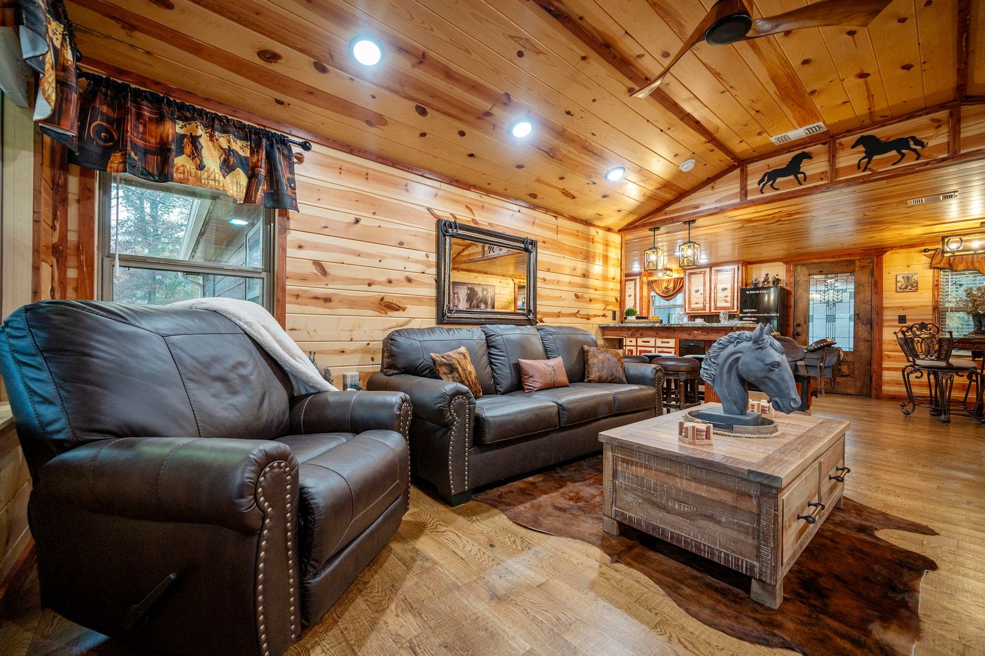 Living room with log walls, dark leather furniture, a horse sculpture on a wooden coffee table, and a cowhide rug.