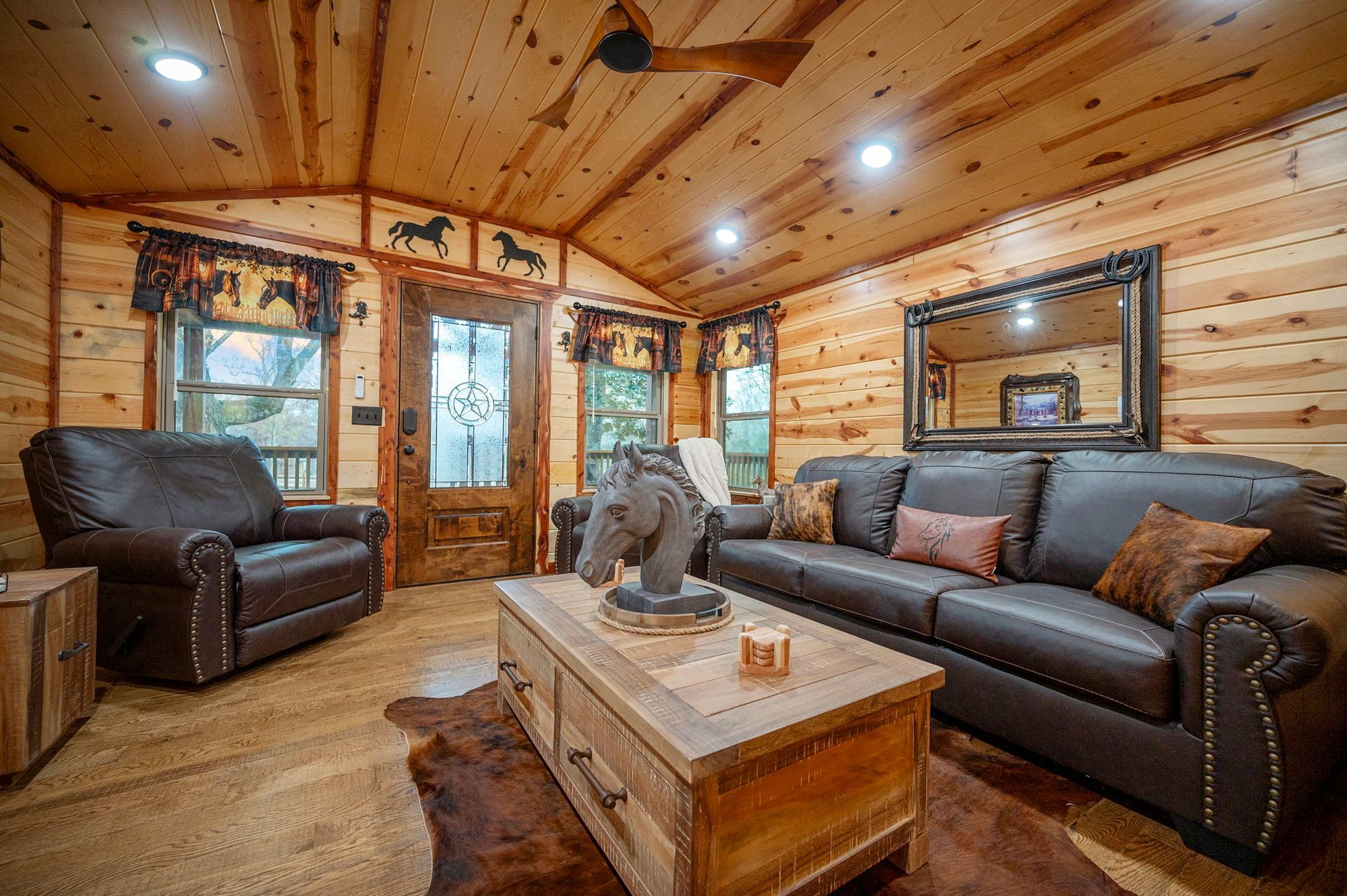 A cozy living room with knotty pine walls, a brown leather sofa and armchair, a coffee table, and a horse-themed decor.