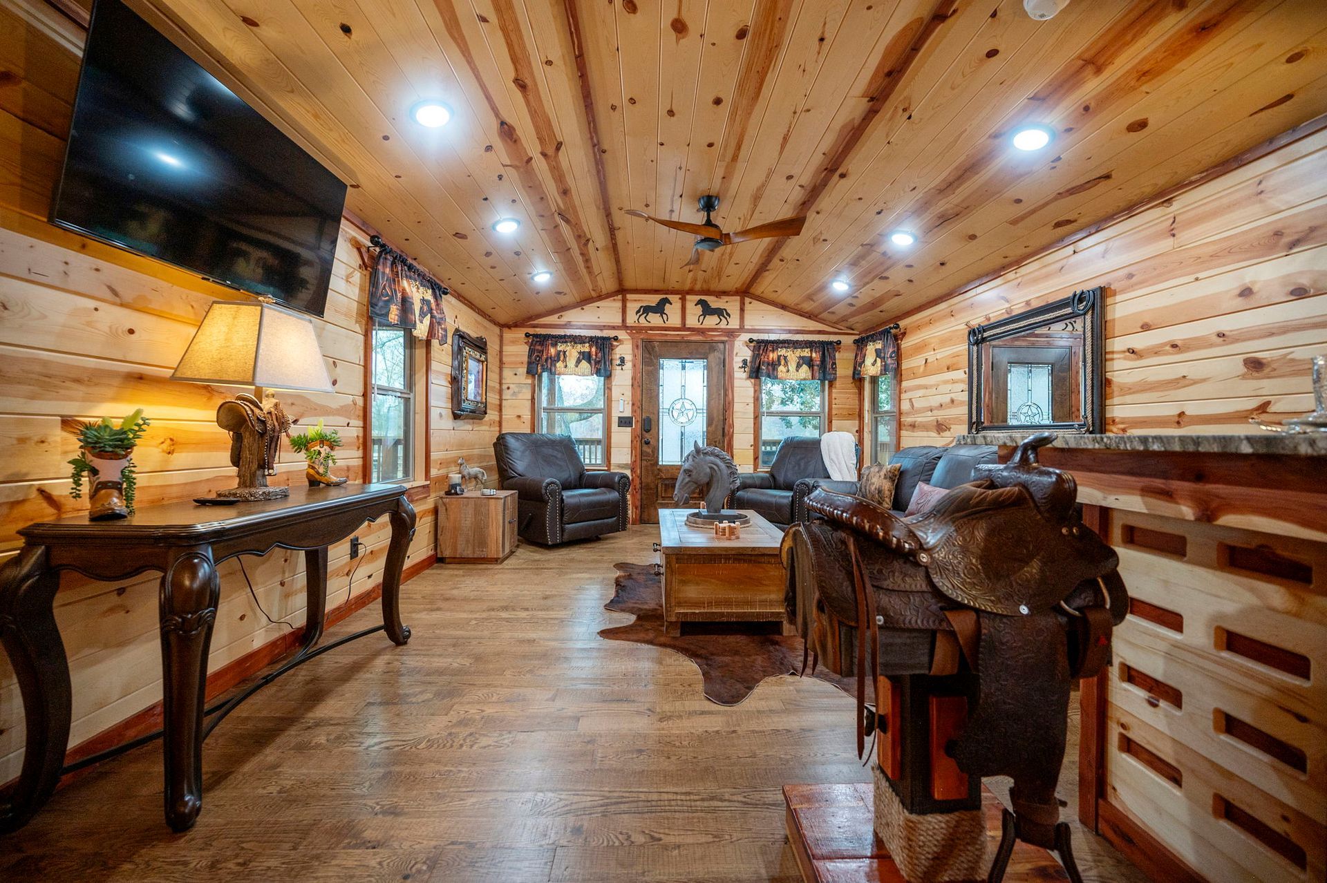 A rustic cabin living room with wood paneled walls, a leather armchair, a central coffee table, and a horse saddle.