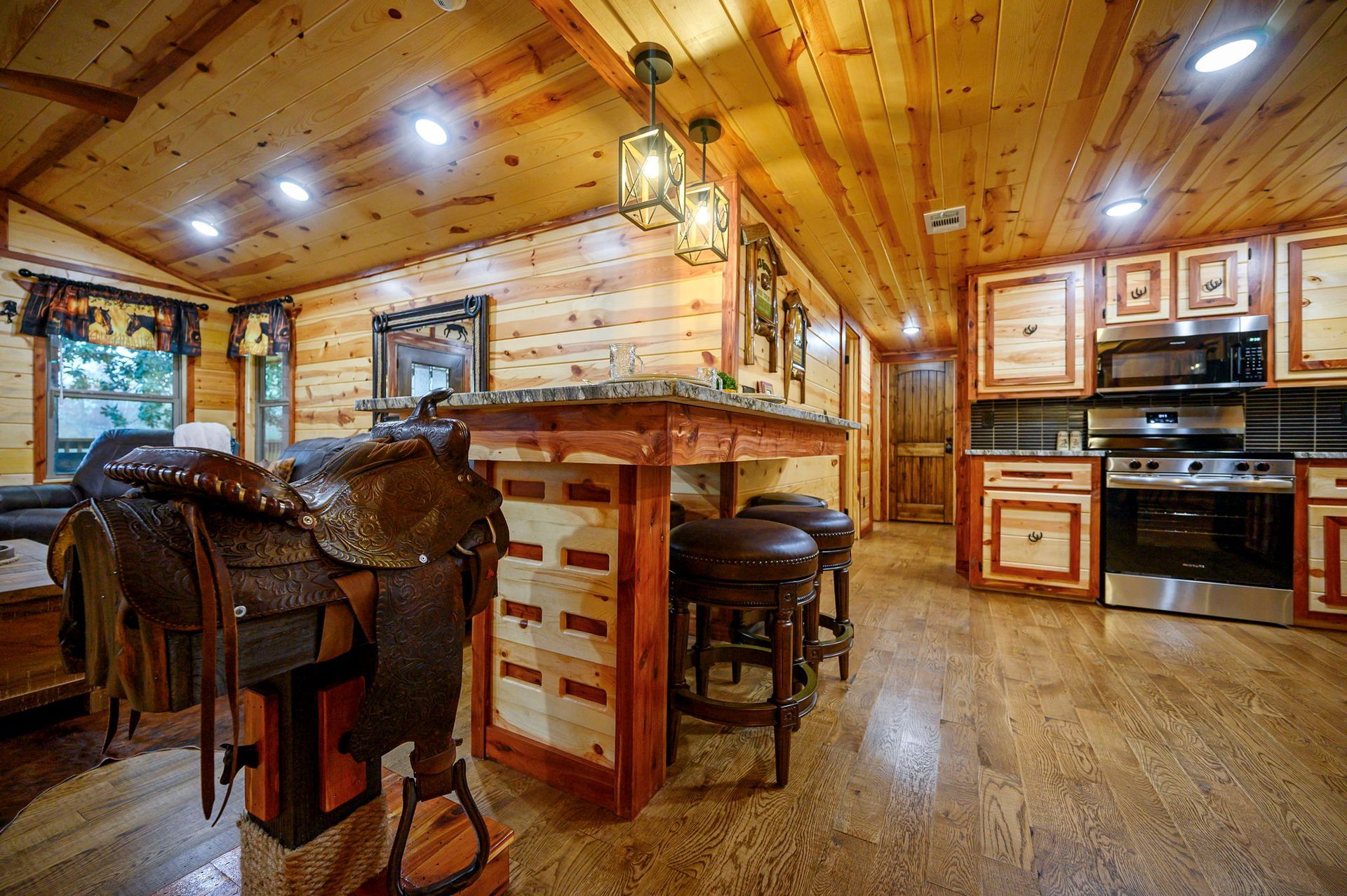 A rustic kitchen with wood-paneled walls and ceilings, featuring a saddle on a stand and a kitchen island with bar stools.