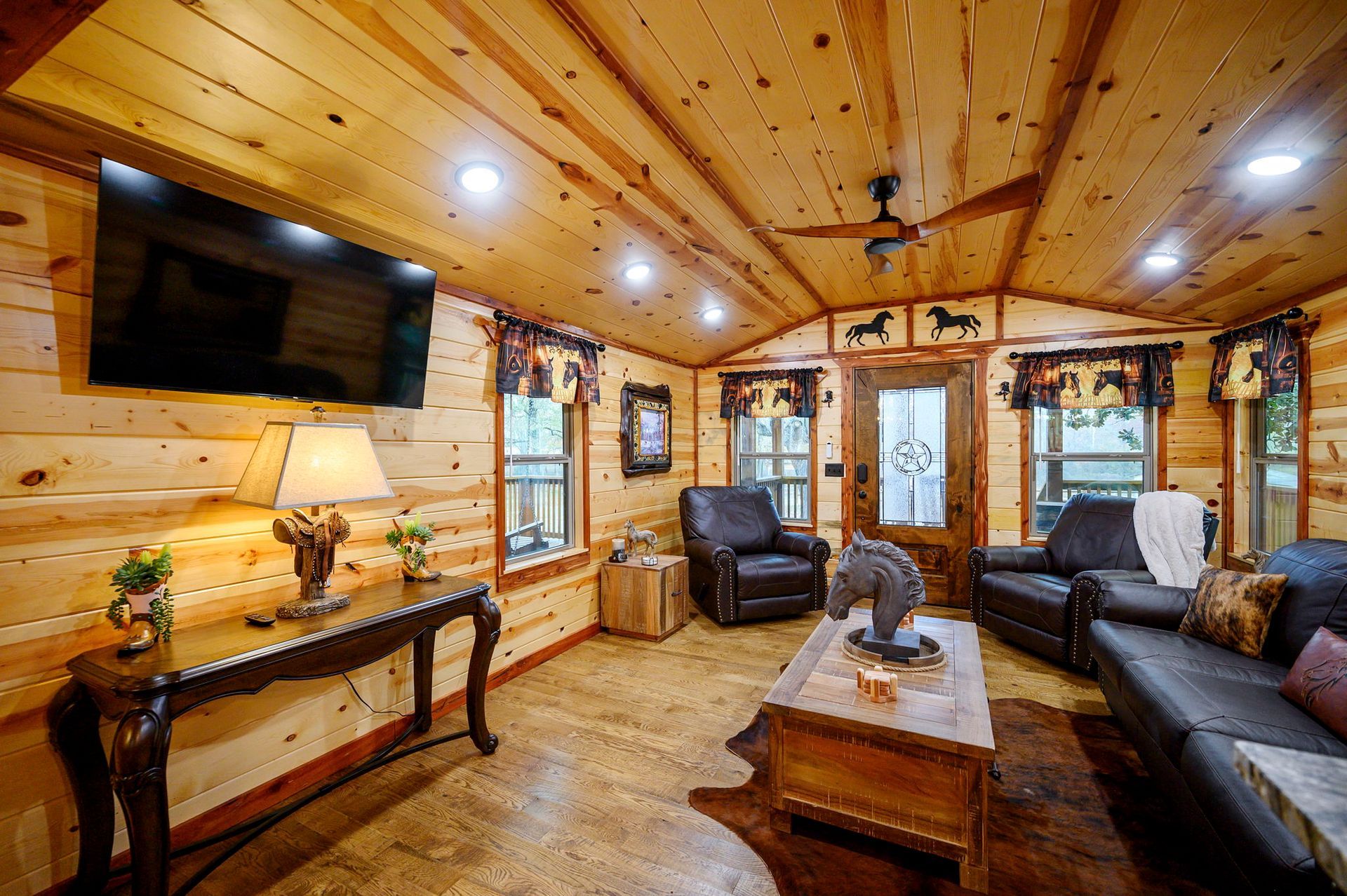 A rustic living room with pine walls and ceiling, leather furniture, a wooden coffee table, and a wall-mounted TV.