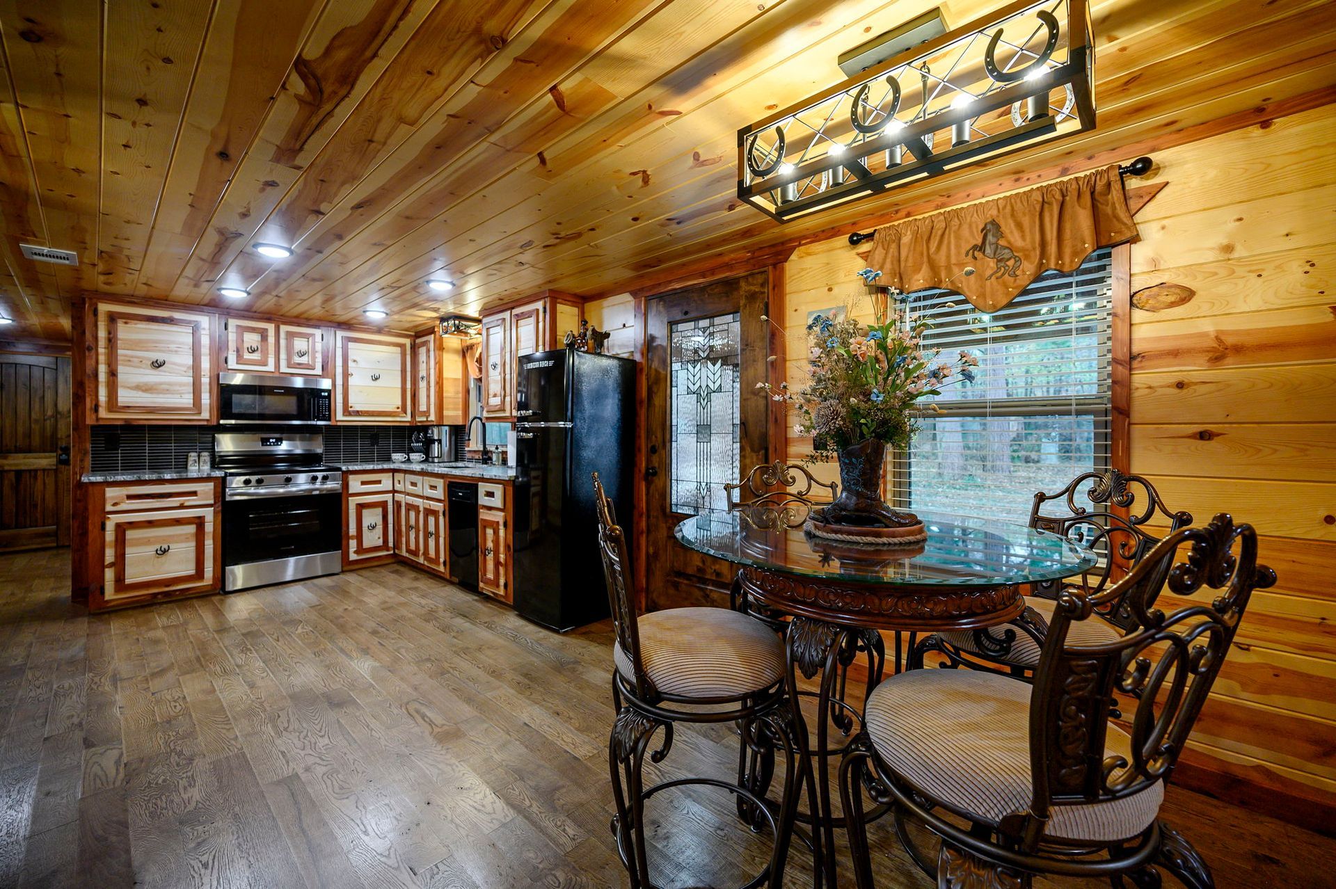 A rustic kitchen with wood-paneled walls and ceilings, stainless steel appliances, and a small glass-topped dining table.
