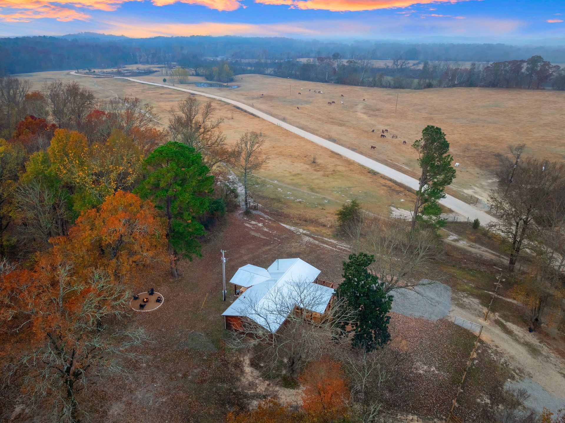 Aerial view of a house with a white roof nestled in a rural landscape with autumn trees and a long, winding dirt road.