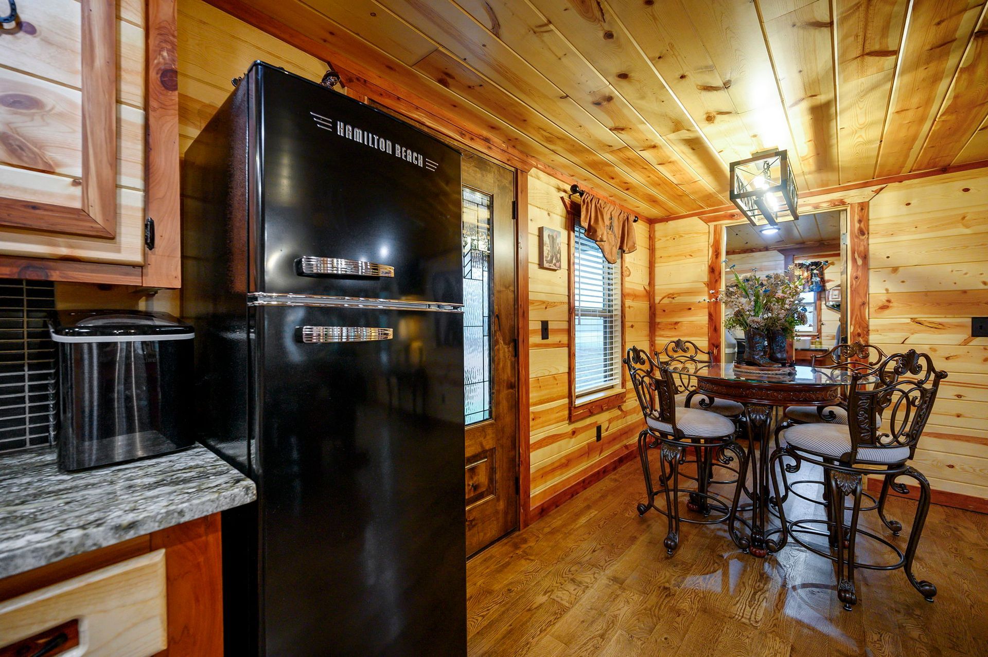 A black refrigerator stands in a rustic wooden kitchen next to a small dining area with a round table and metal chairs.