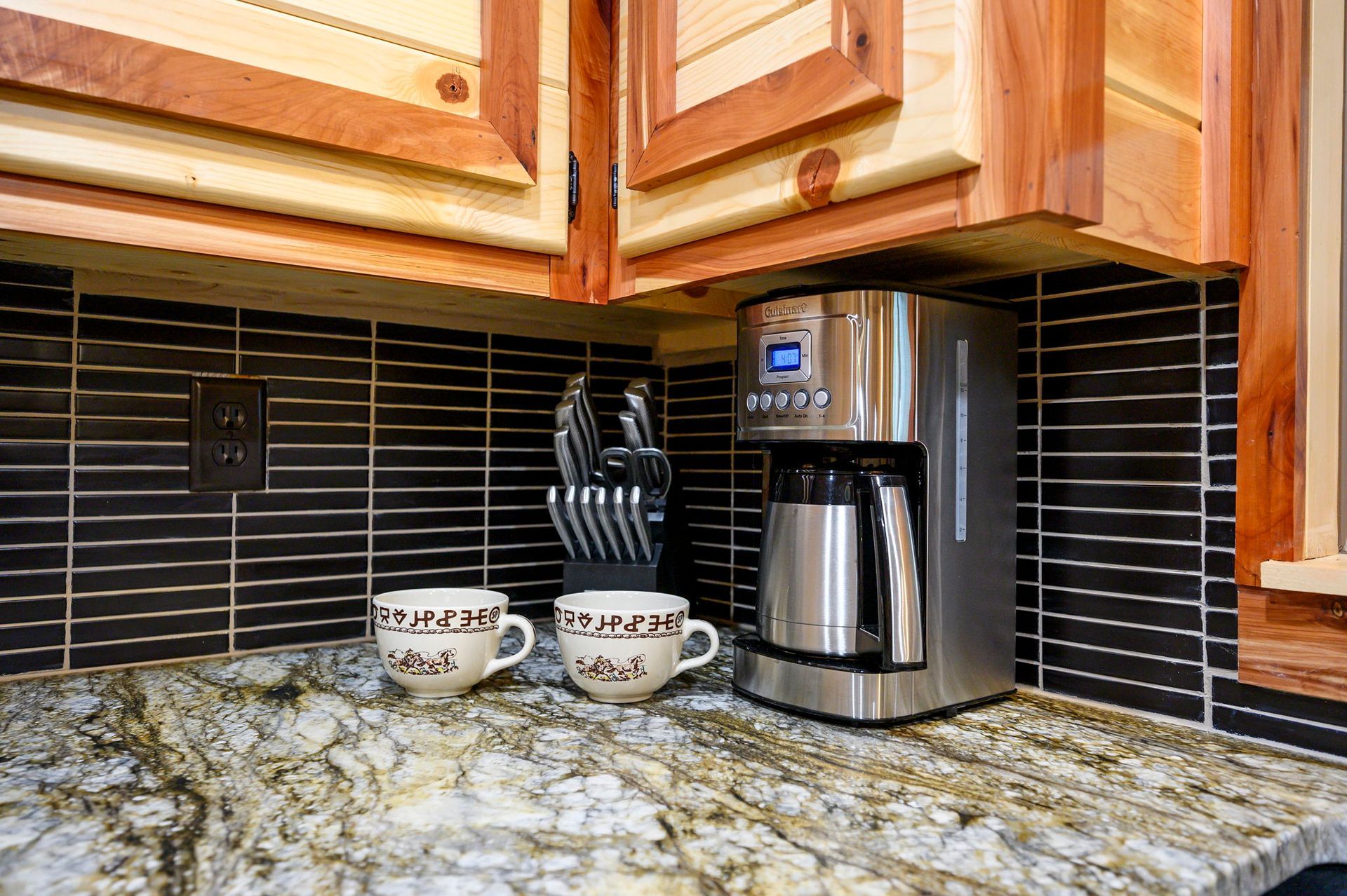 A stainless steel coffee maker sits on a granite countertop next to two white mugs and a knife block against black tiles.