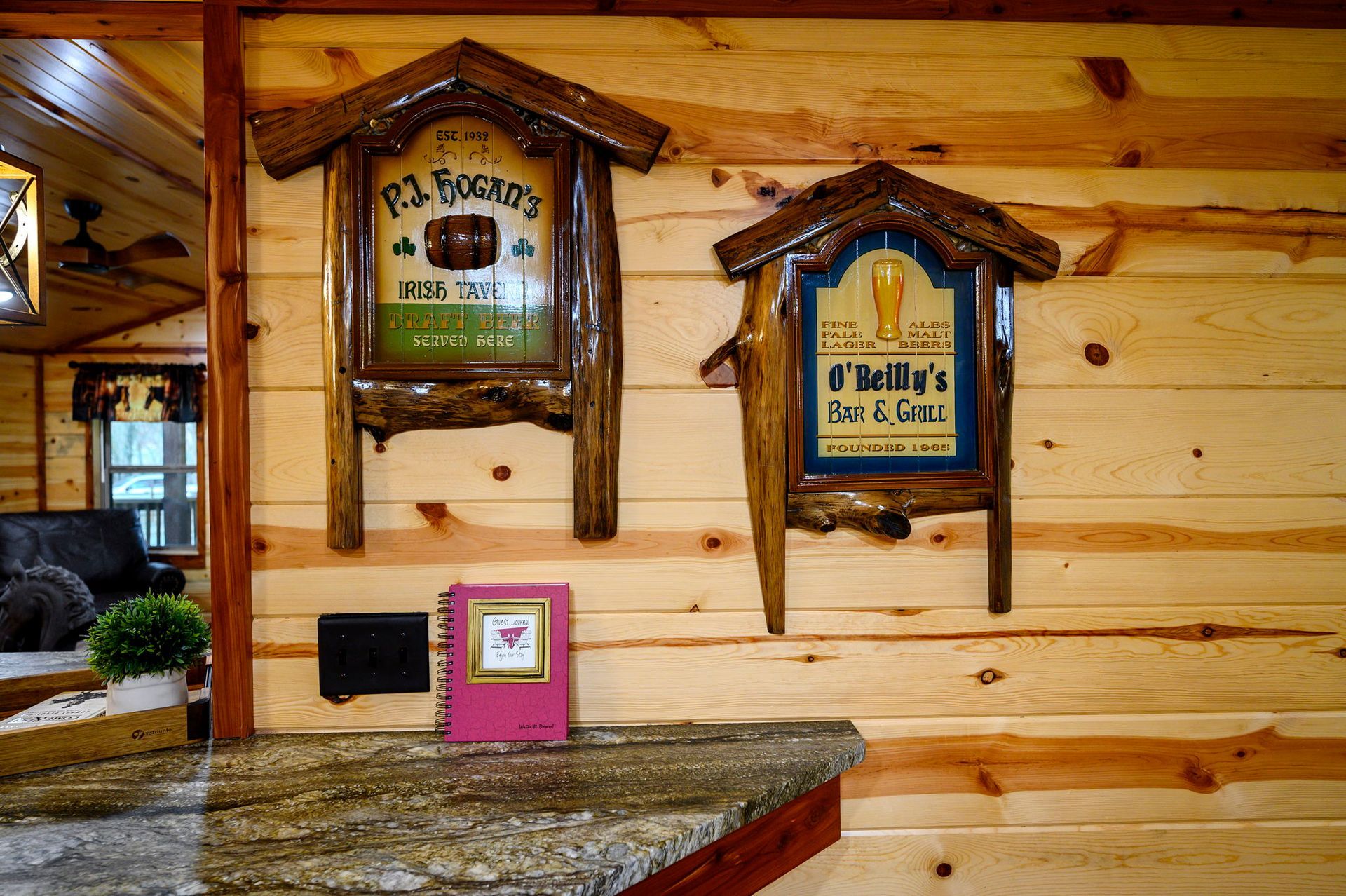 Two rustic wooden signs and a small pink book sit on a stone counter against a knotty pine log wall.