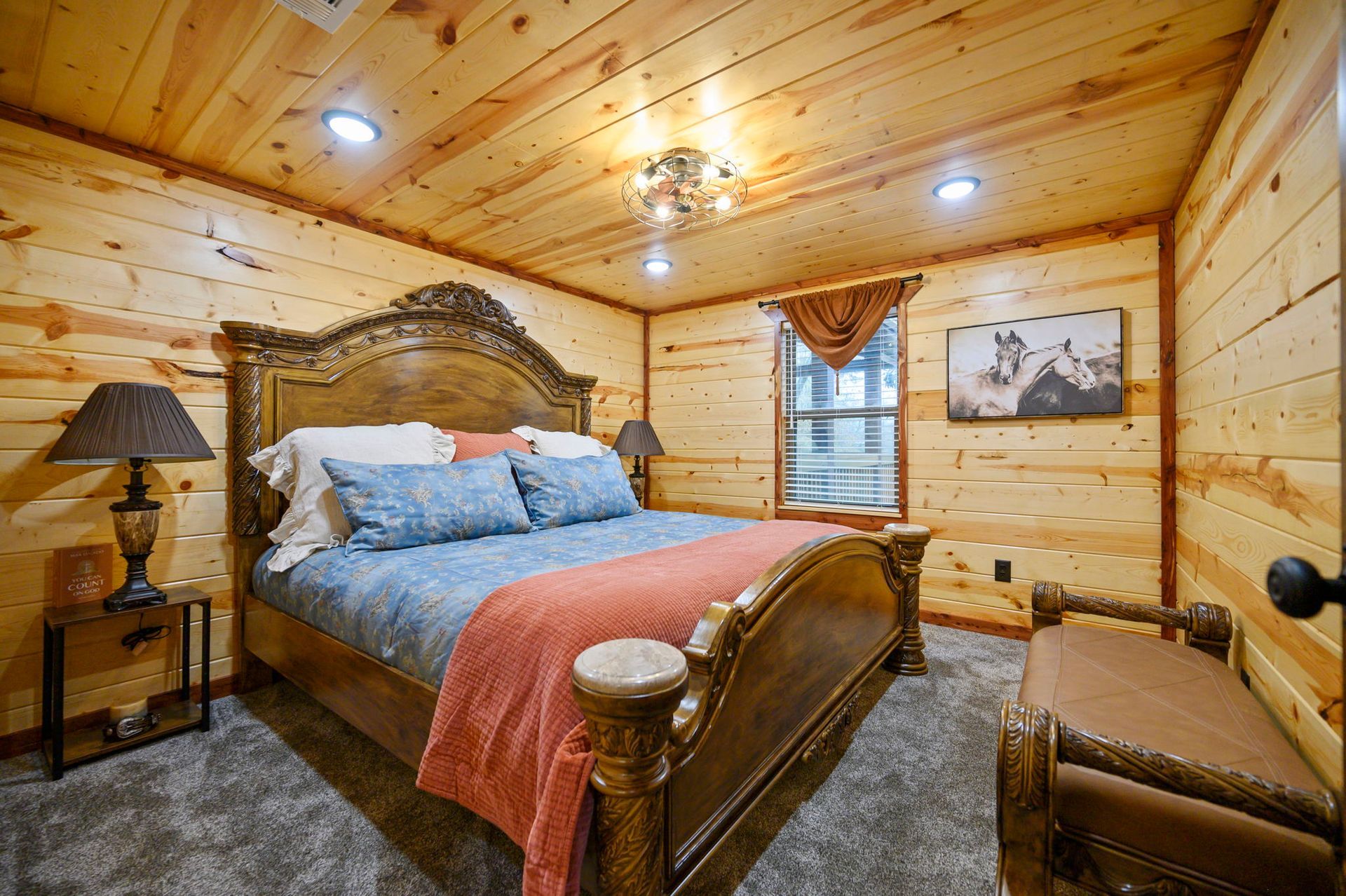 A rustic bedroom features a wooden bed with blue bedding, a coral throw, matching lamps, and a wooden bench on carpet.