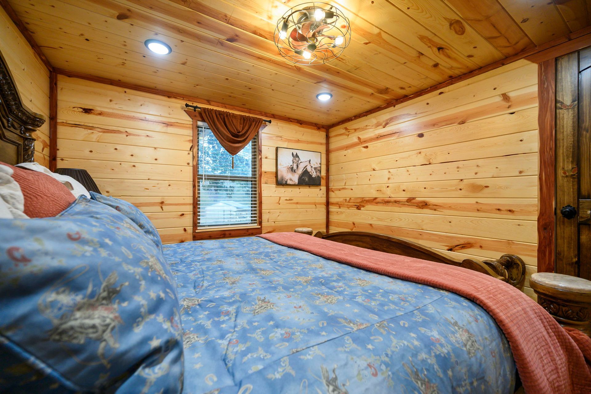 A bedroom with knotty pine walls and ceiling, a bed with a blue patterned comforter, and a window with a valance.