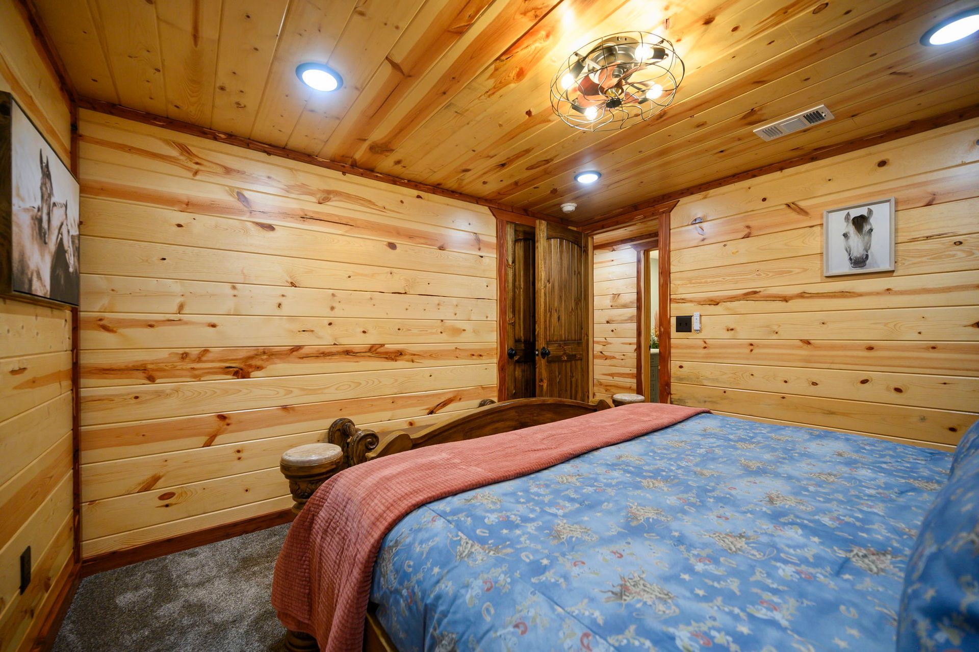 A rustic bedroom with wooden walls, a bed featuring a blue floral quilt and pink throw, and a decorative ceiling light.