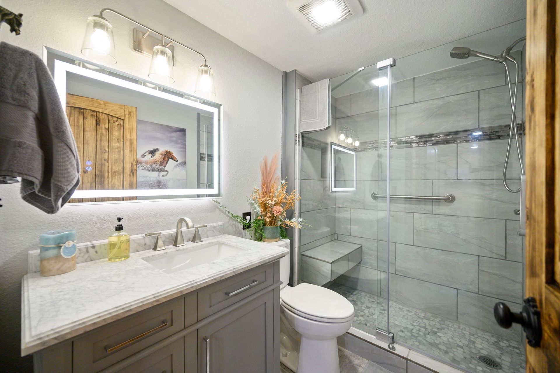A modern bathroom with a gray vanity, marble countertop, backlit mirror, and a glass-enclosed shower with tiled walls.