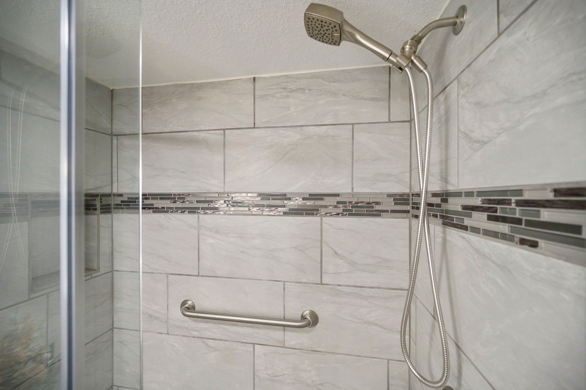 A walk-in shower with light grey tile, a decorative horizontal mosaic strip, a handheld showerhead, and a grab bar.