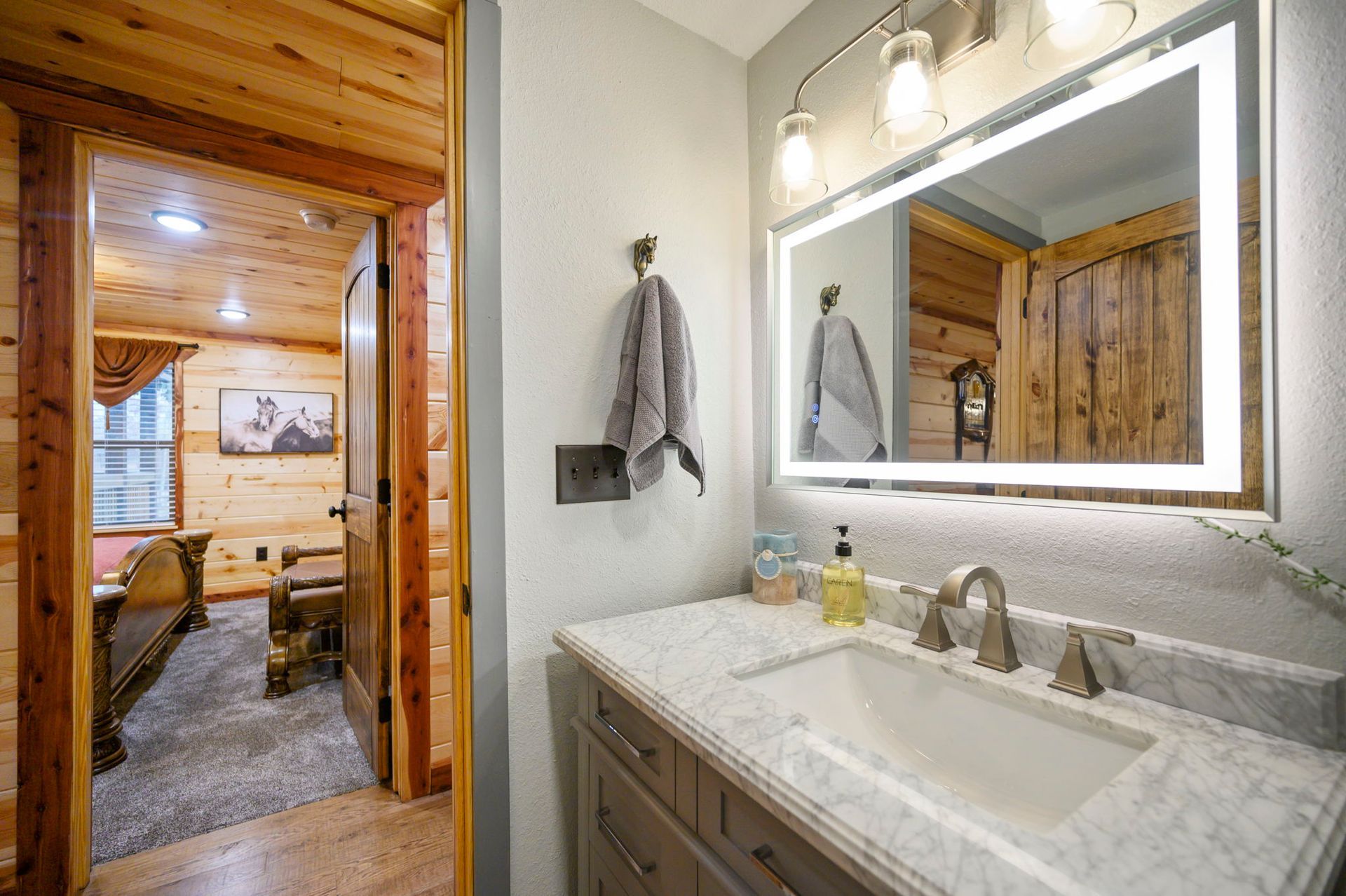 A bathroom vanity with a marble countertop, silver faucet, and an illuminated mirror, next to a doorway into a bedroom.