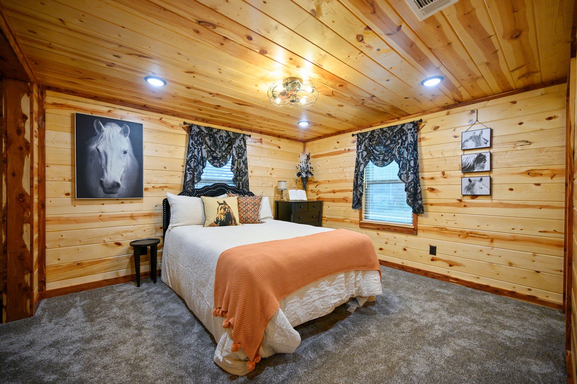 A rustic bedroom with wood-paneled walls, a bed with a white comforter, an orange throw, and framed horse-themed art.