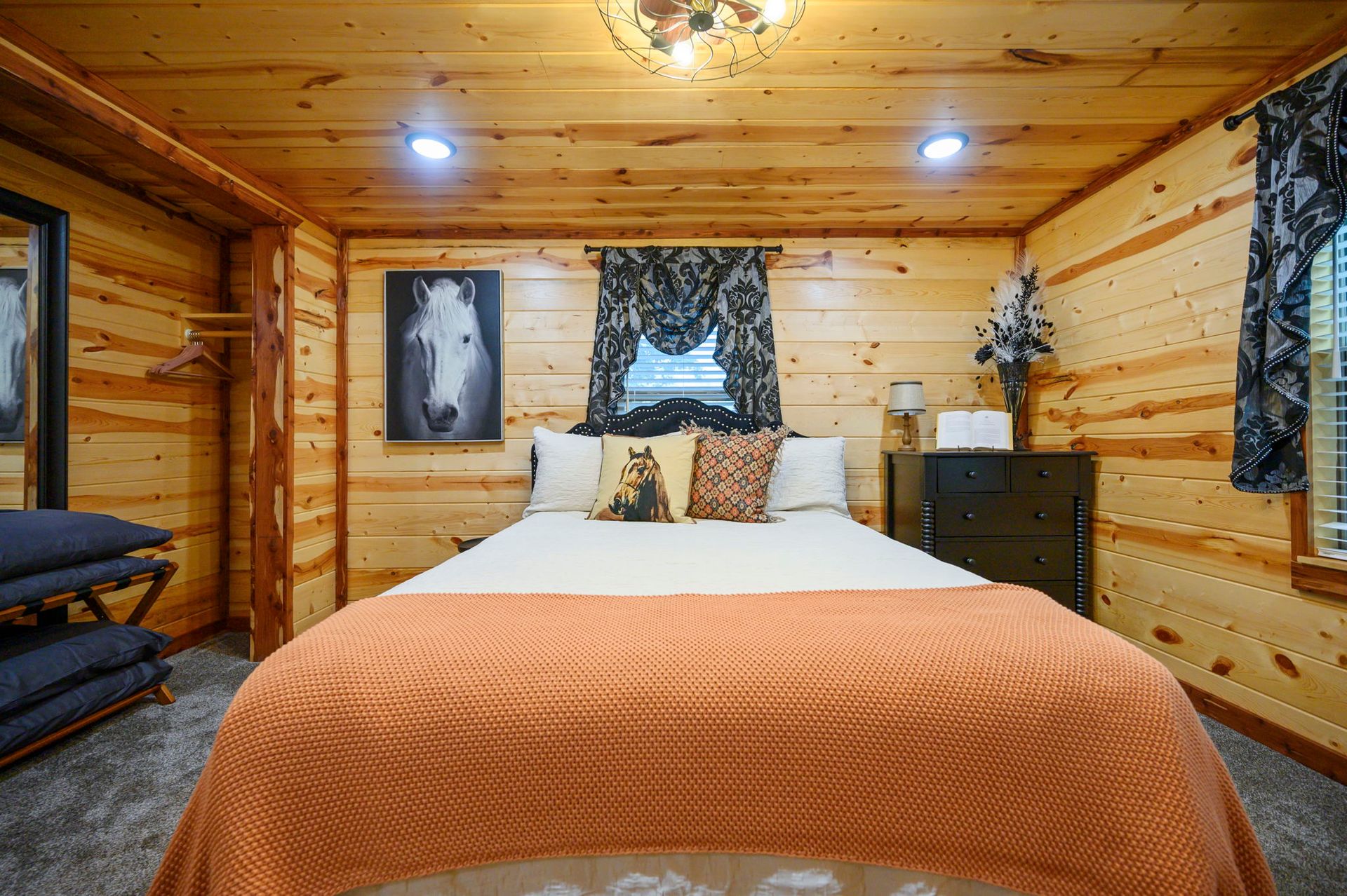 A rustic bedroom with wood-paneled walls, a bed with an orange knit blanket, and a white horse portrait on the wall.