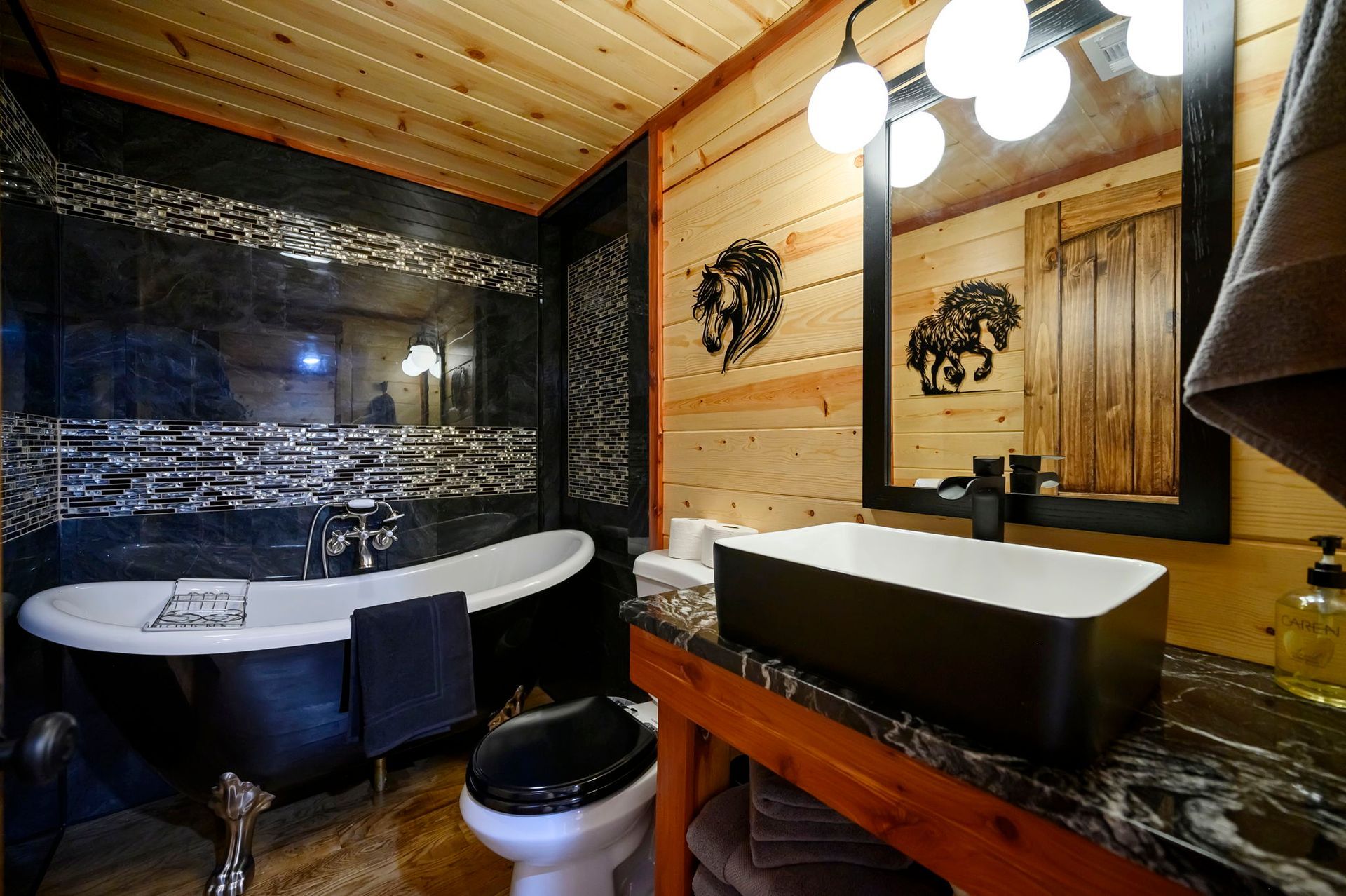 A rustic bathroom with a clawfoot tub, a dark vessel sink on a wooden vanity, and pine wood walls with black decor.