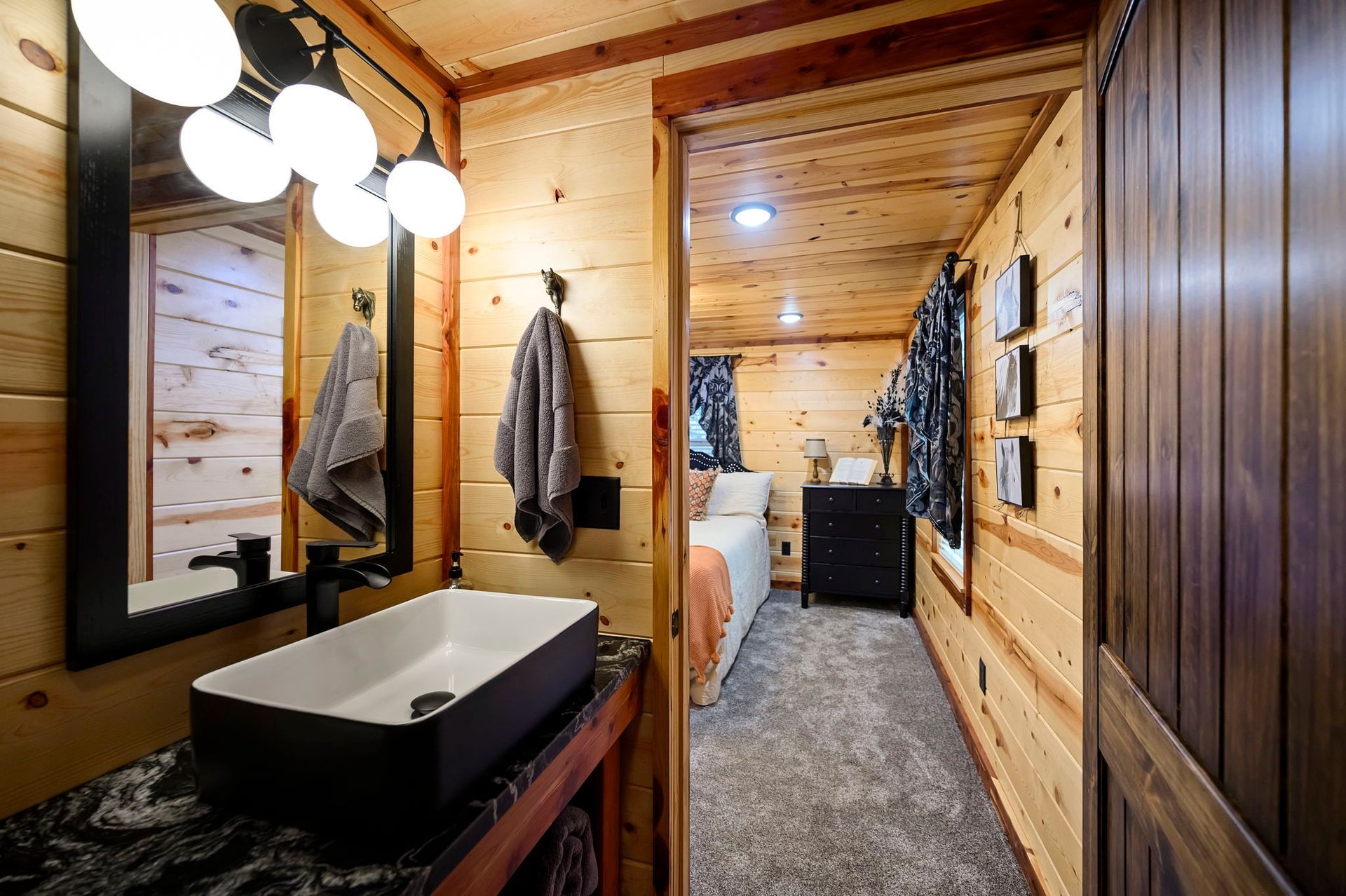 A rustic bathroom with a white vessel sink on a dark vanity opens into a bedroom with wood-paneled walls and a bed.