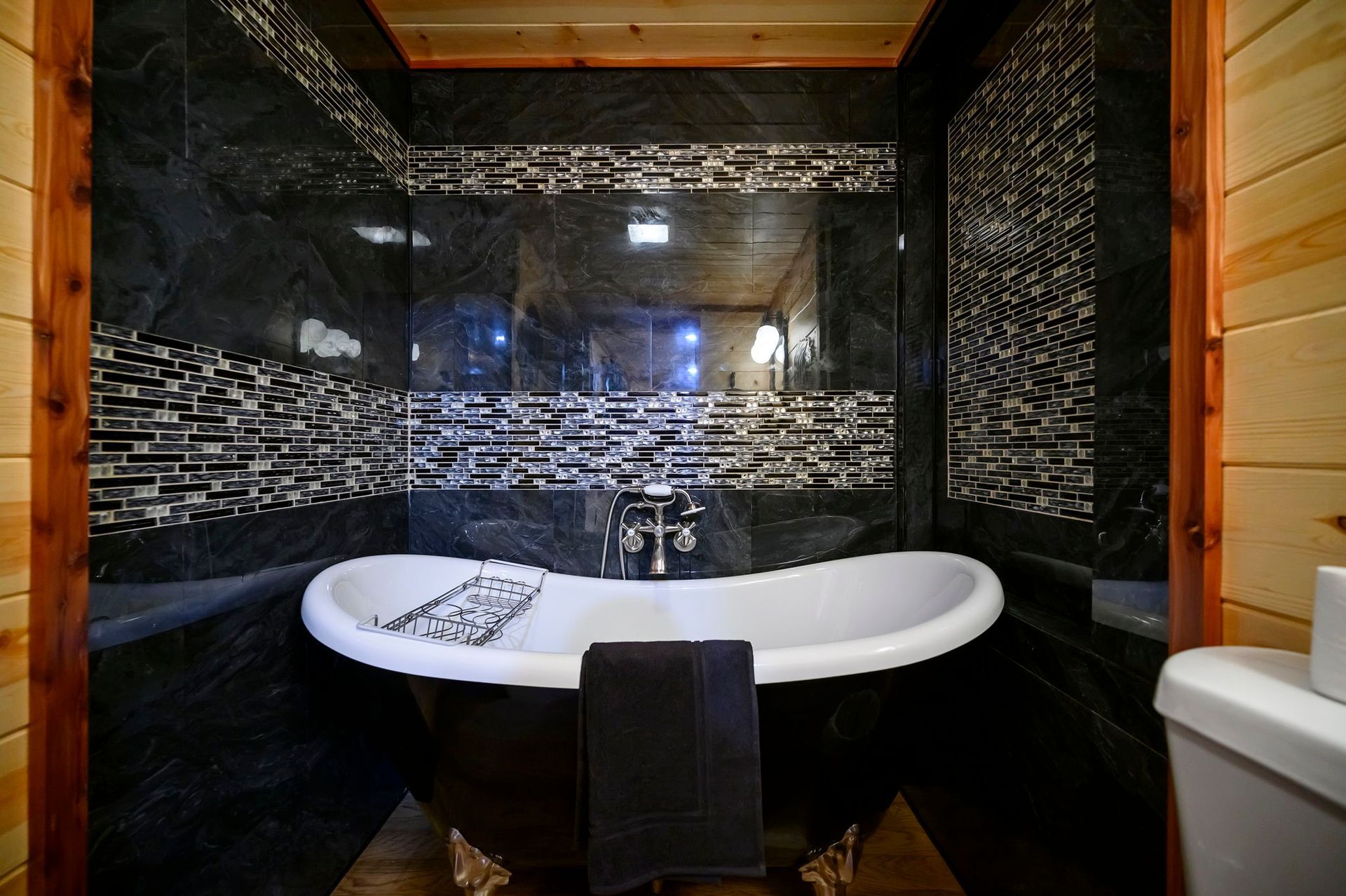 A white clawfoot bathtub with a black exterior centered in a bathroom featuring black tile walls and wood paneling.