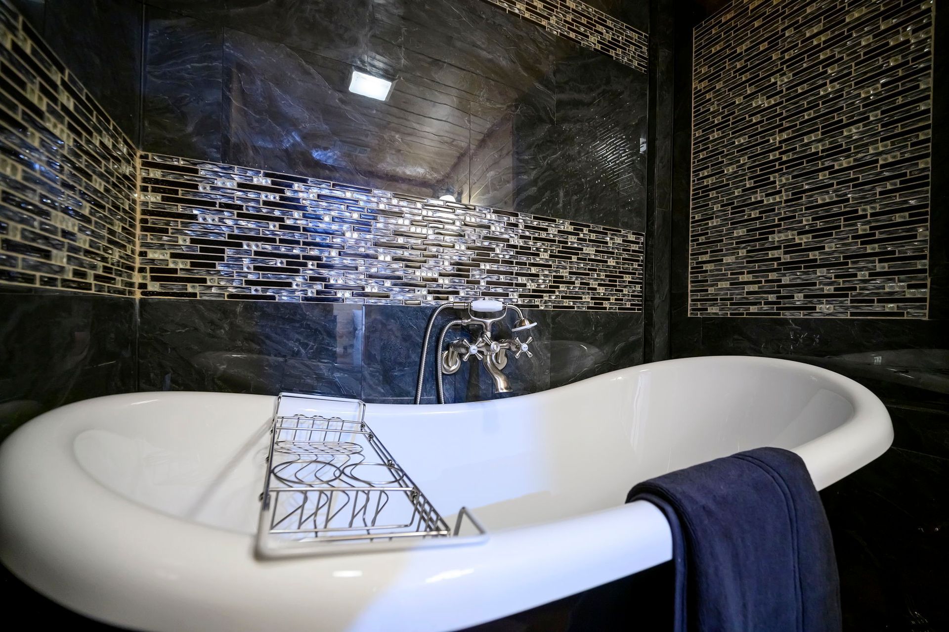 A white claw-foot bathtub sits against a wall with black and gold mosaic tiles. A dark blue towel hangs over the edge.