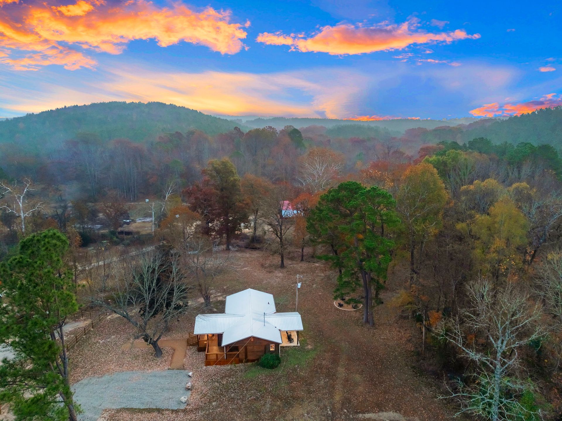 An aerial view of a cabin nestled in a wooded landscape at sunset, with autumn-colored trees under a vivid orange sky.