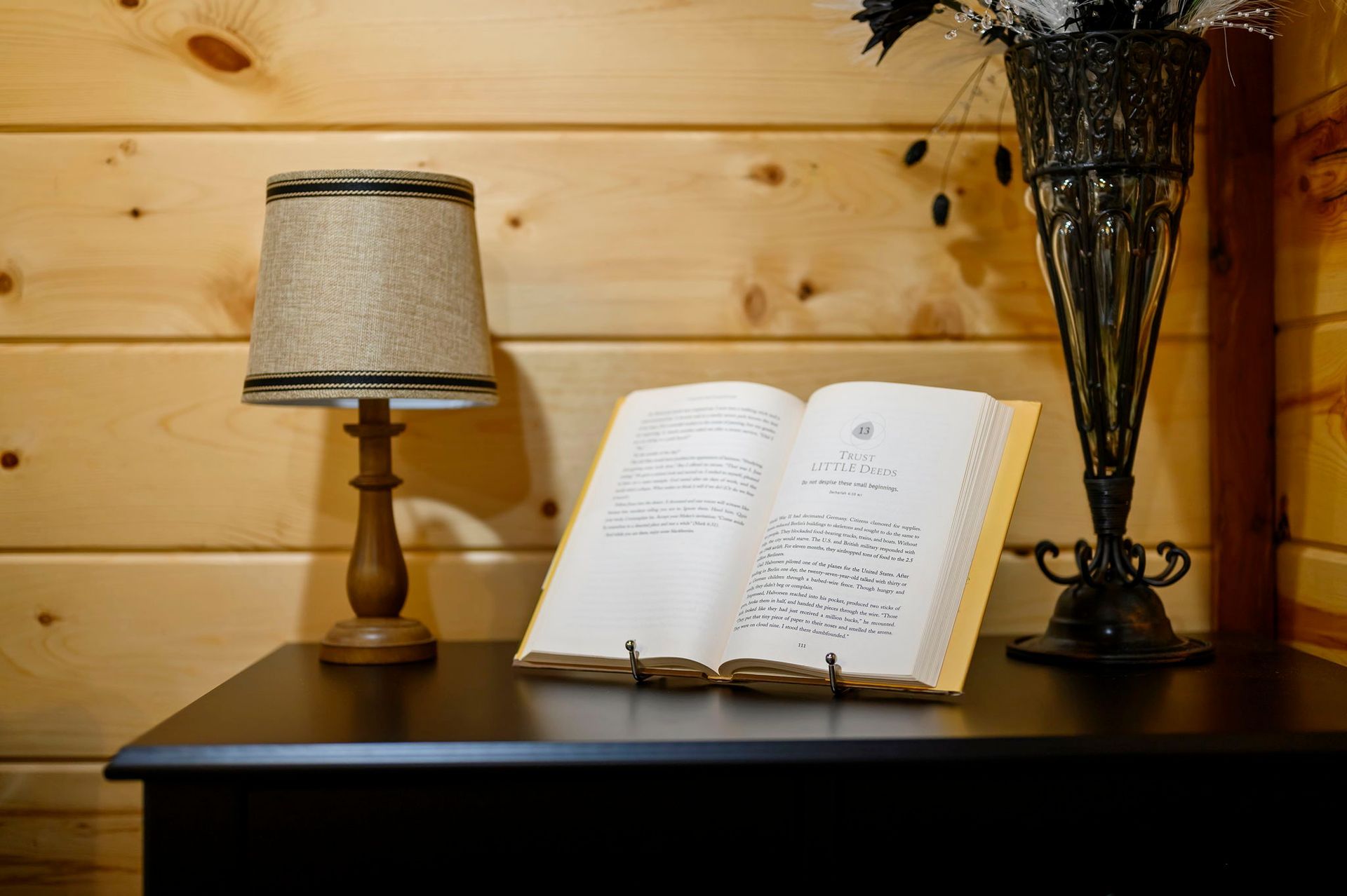 An open book on a wooden stand sits on a dark table next to a small lamp and a decorative vase against a wood-paneled wall.