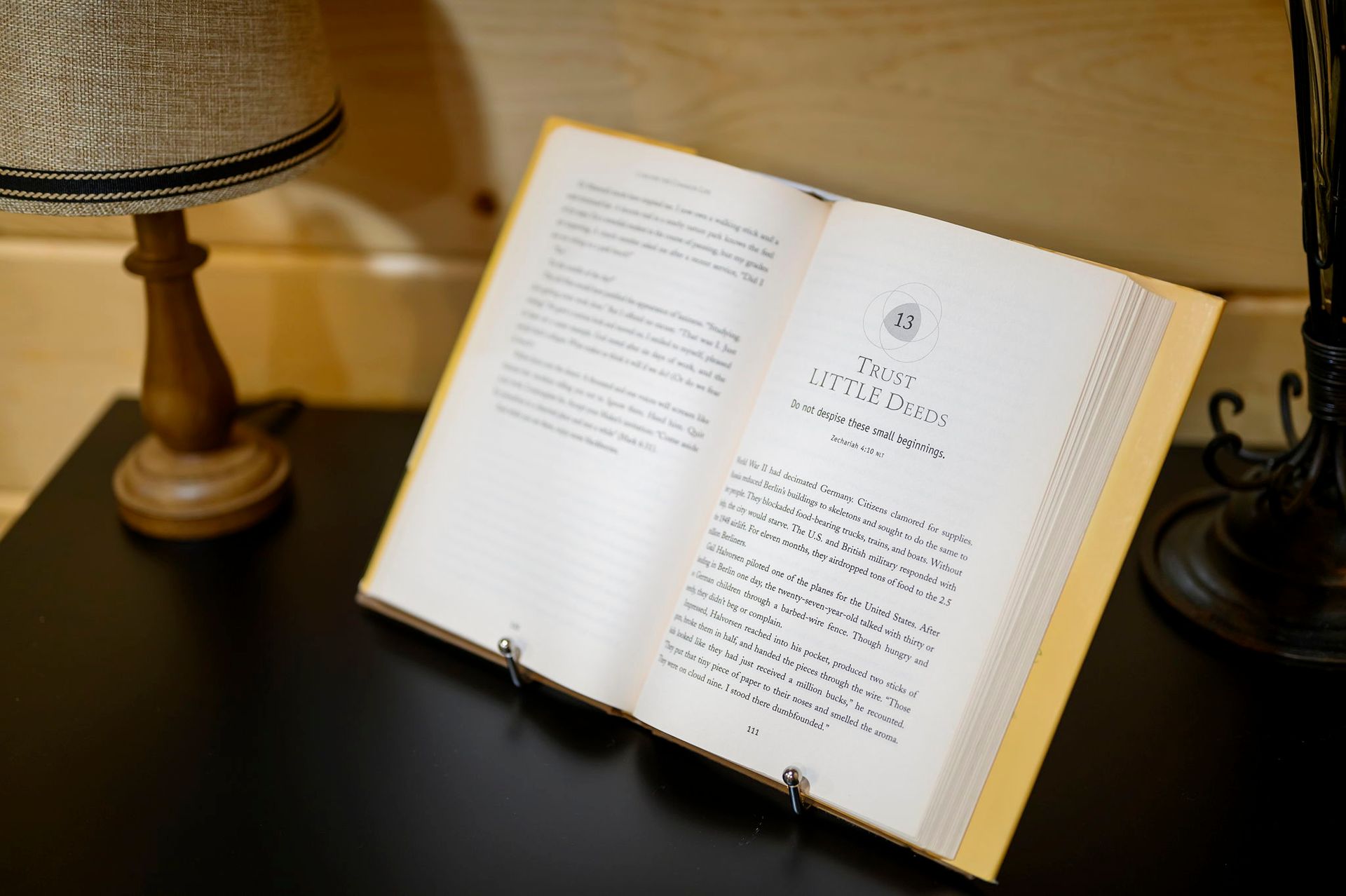 An open book rests on a stand on a black table between two lamps in a room with light-colored wooden walls.