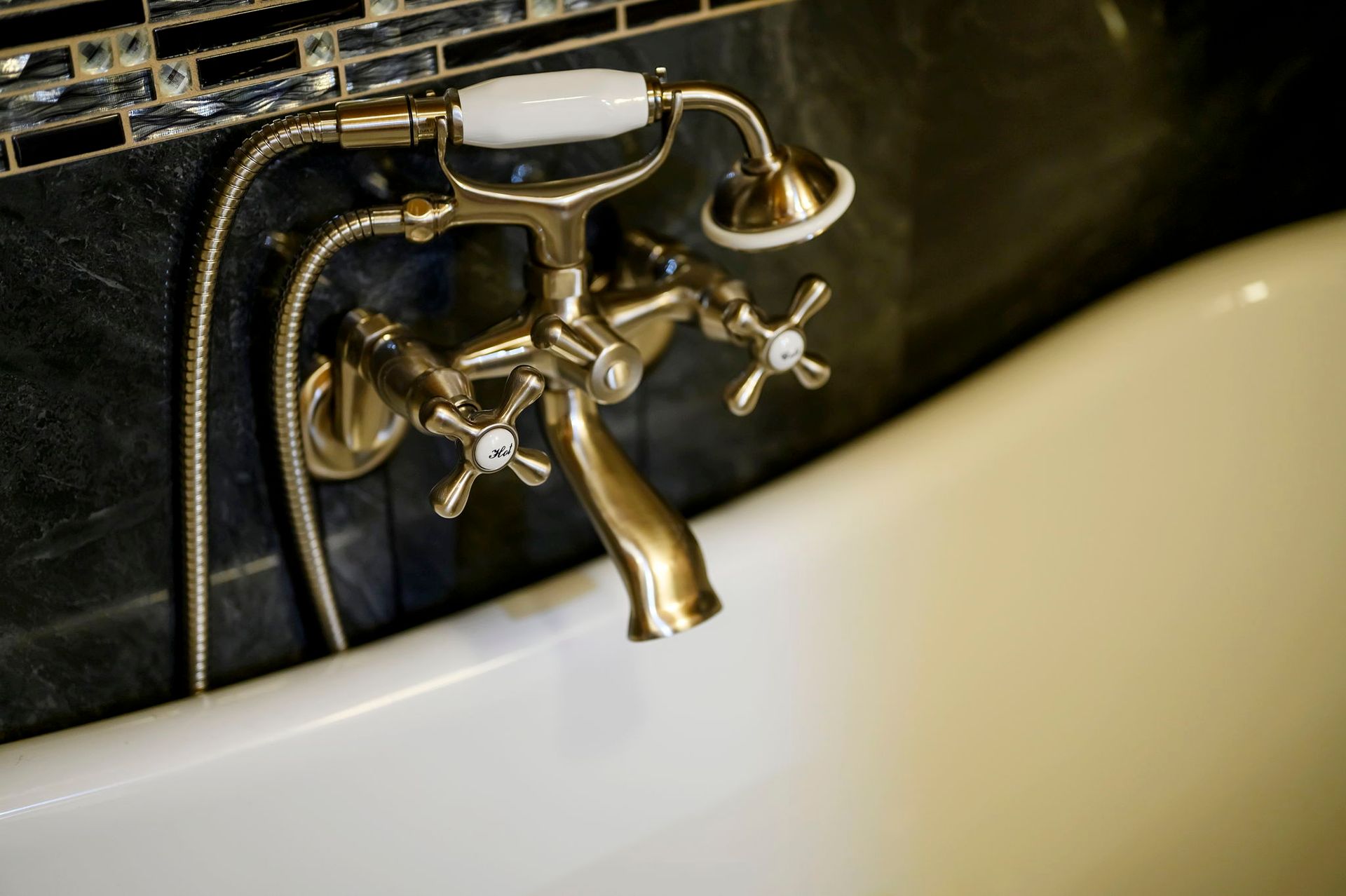 A vintage-style brass bathtub faucet with a handheld sprayer attached, mounted on a dark tiled wall above a white tub.