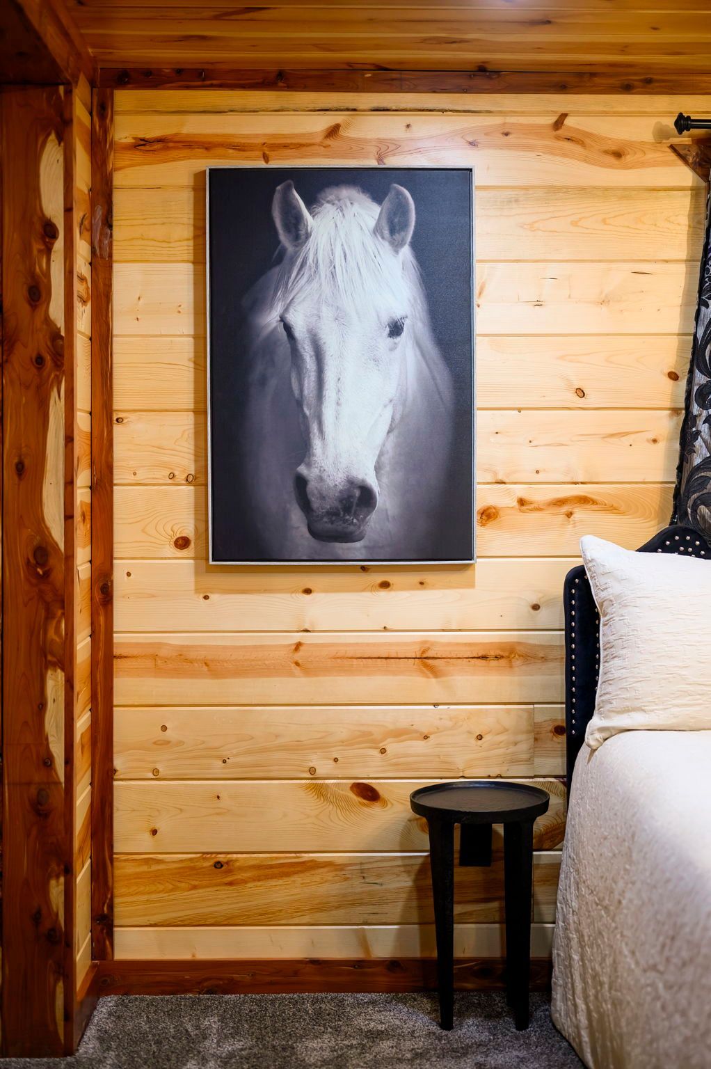 Black and white portrait of a horse hanging on a wood-paneled wall above a small side table next to a bed.