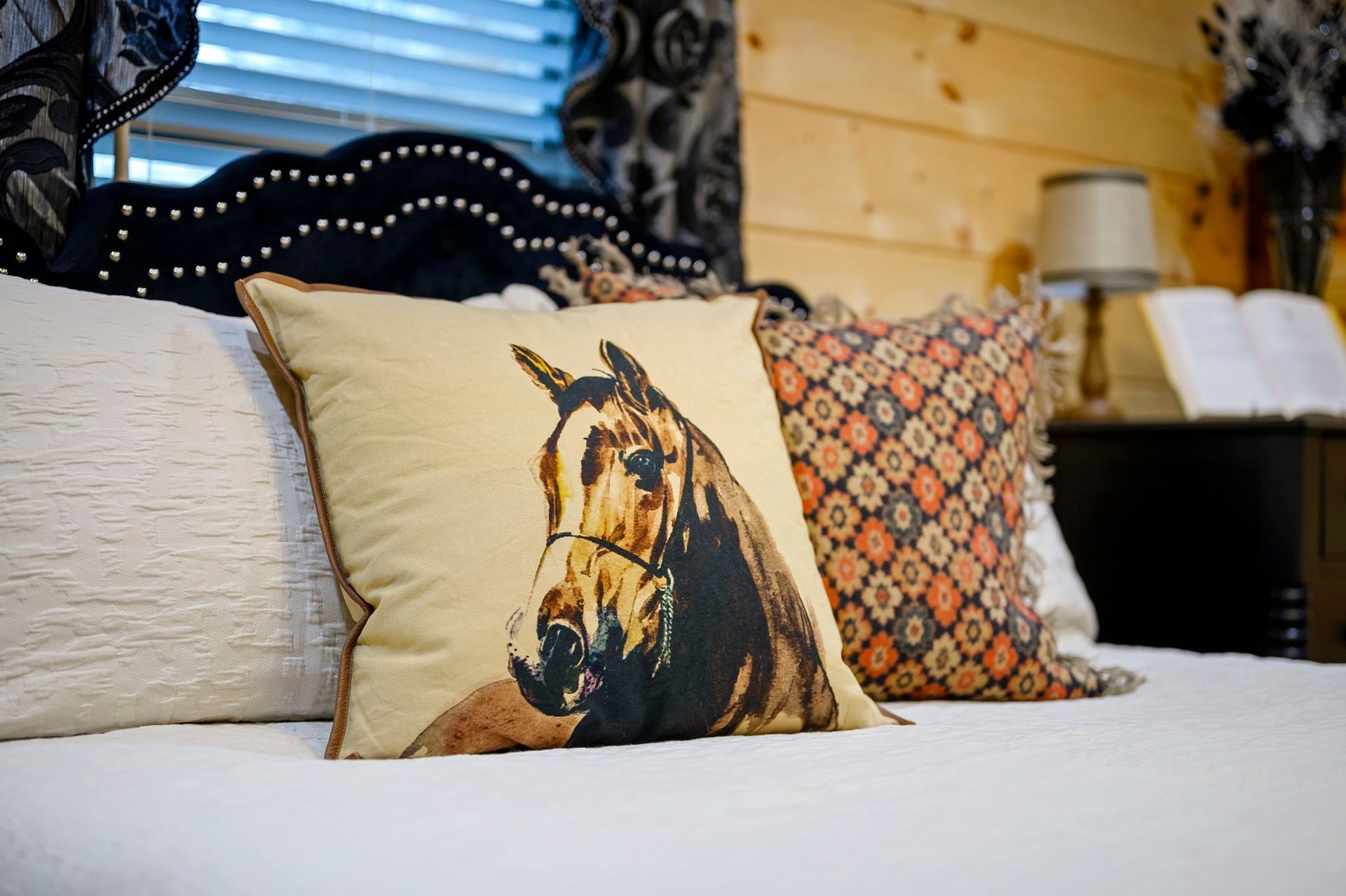 A decorative pillow with a horse print sits on a bed with a white quilt, near a patterned pillow and wooden wall.