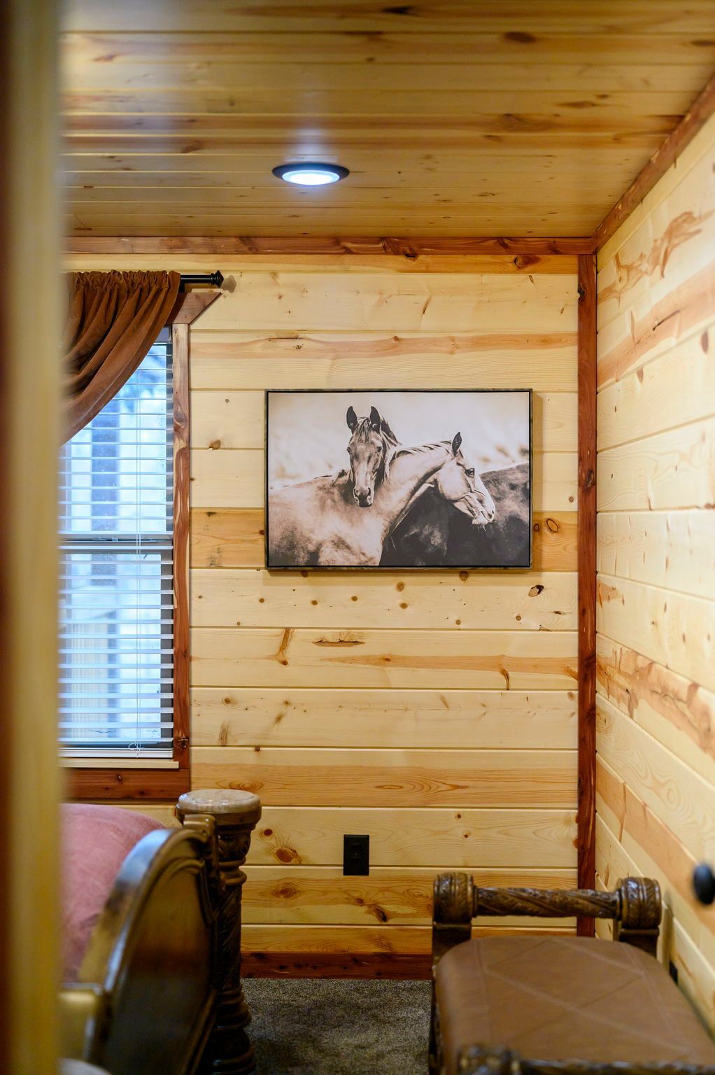 A bedroom with wood-paneled walls features a horse print art piece, a window with curtains, and a brown leather bench.