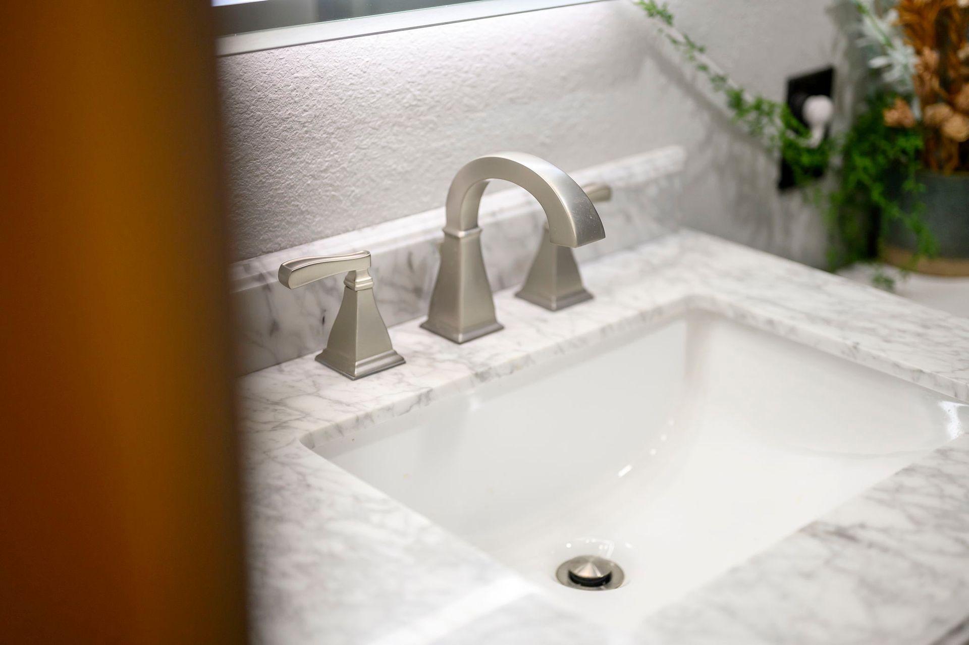 A sleek, brushed-nickel faucet with two side handles mounted on a white and grey marble bathroom vanity countertop.
