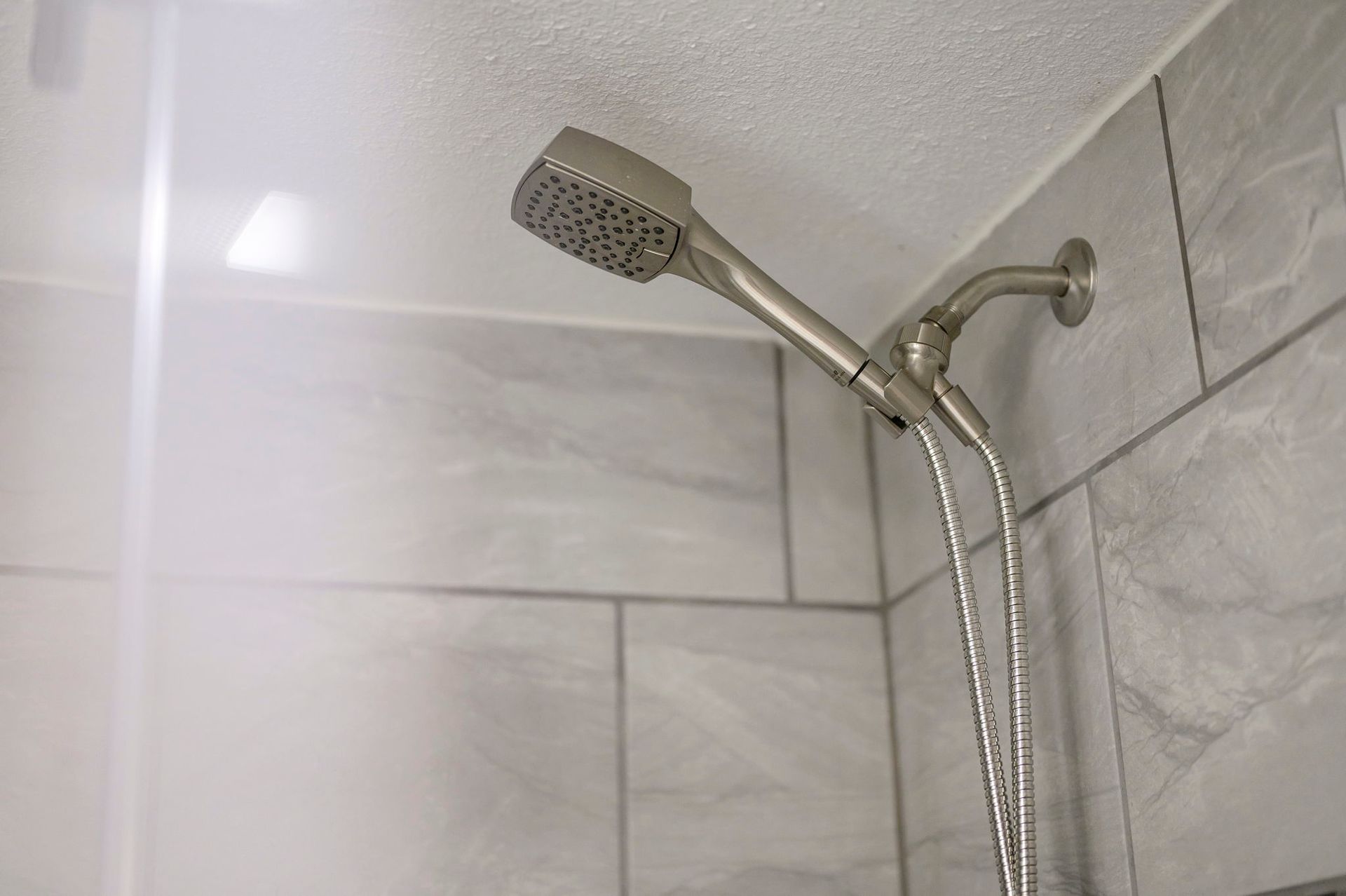 A nickel-finish shower head mounted on a grey tiled wall with a handheld hose attachment.