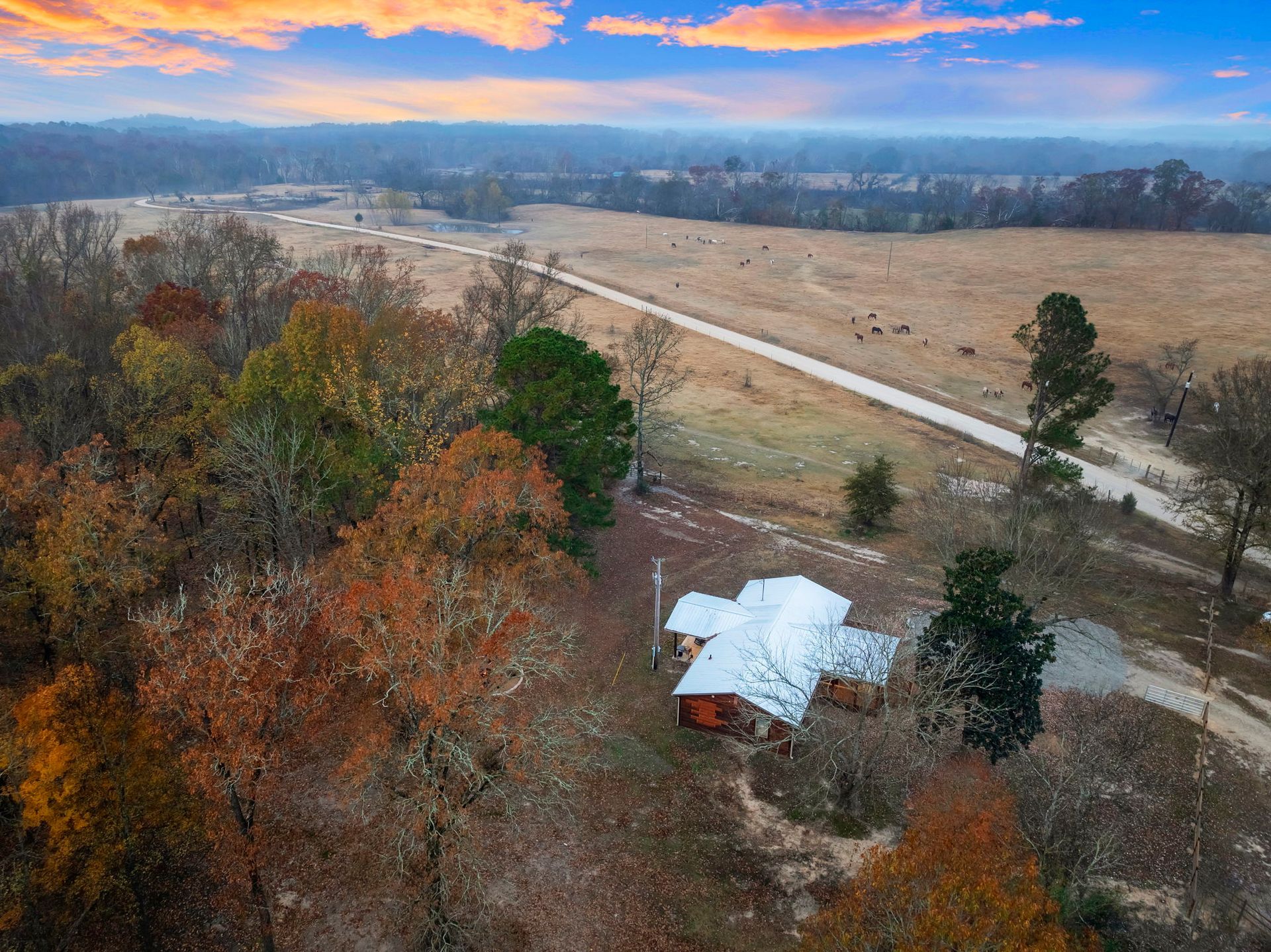An aerial view of a cabin with a tin roof nestled in a rural landscape of autumn trees and a vast, dry field.