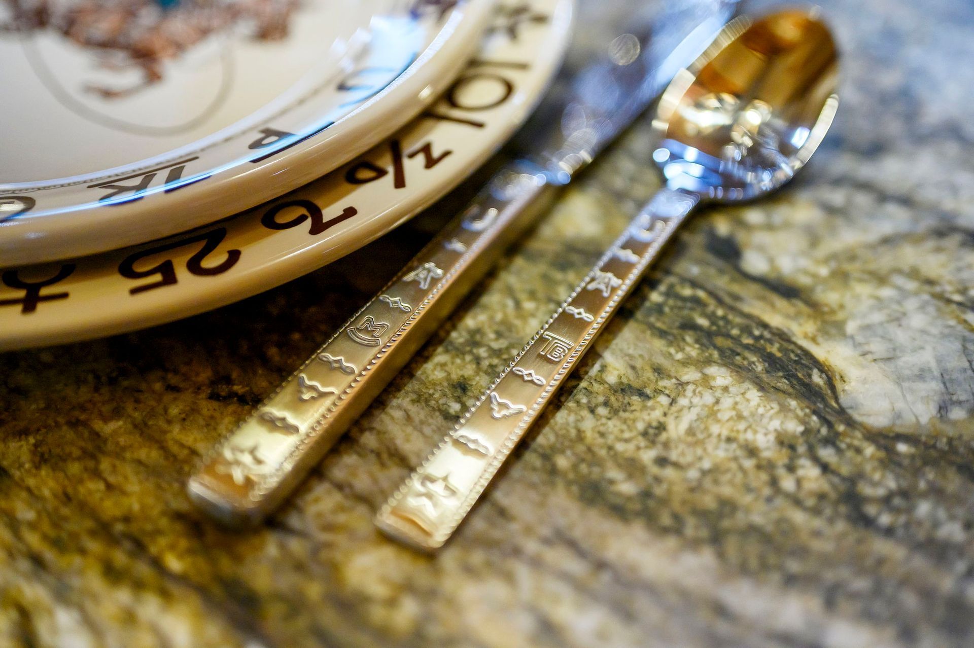 A gold-colored spoon and knife with embossed patterns resting on a textured green surface beside a patterned plate.