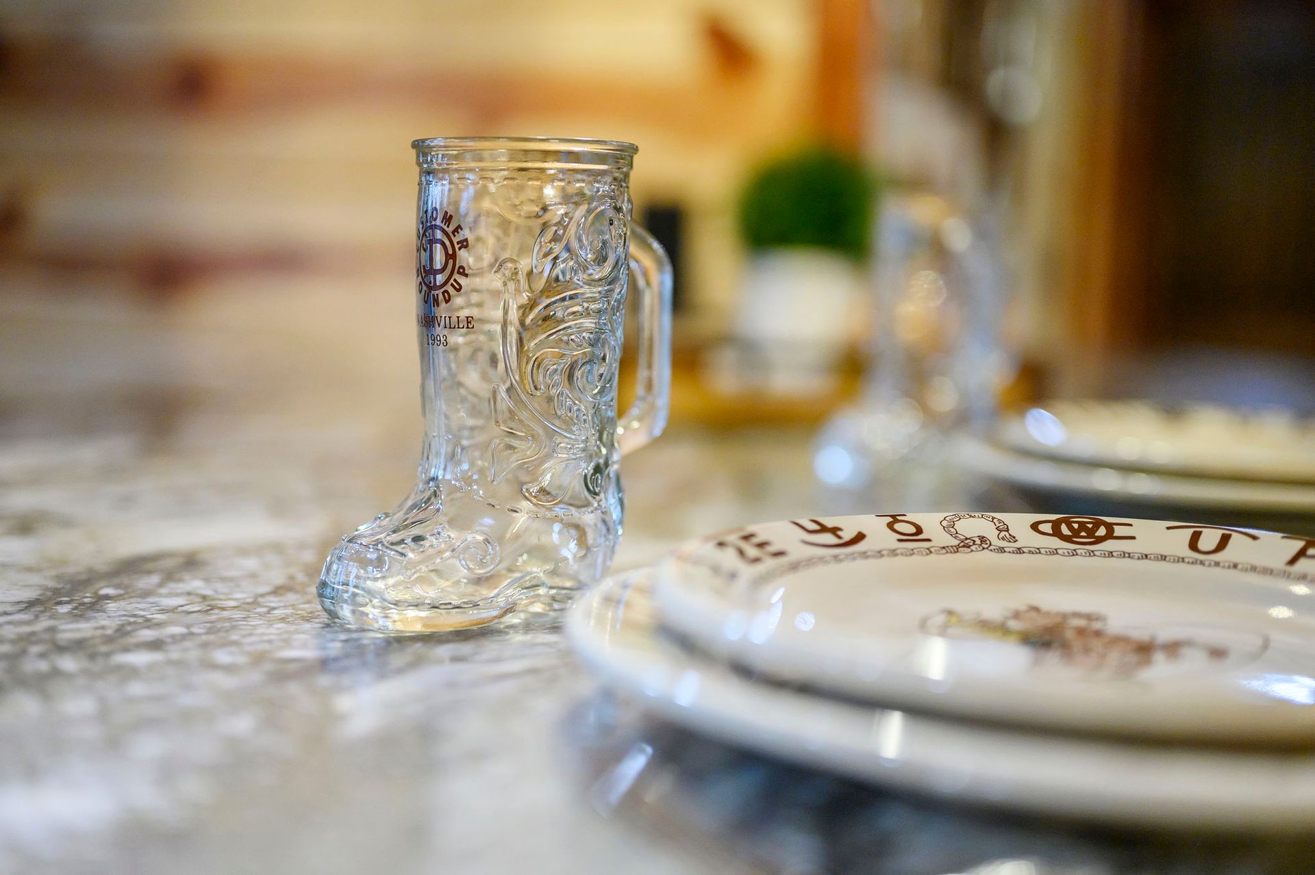 A clear, glass boot-shaped mug stands on a marble surface next to a stack of patterned plates.
