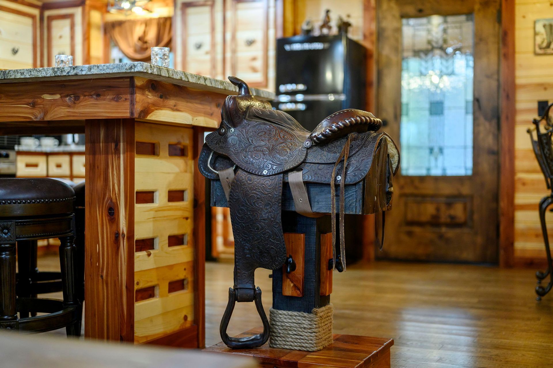 A dark leather western saddle sits on a wooden stand in a rustic, wood-paneled kitchen.