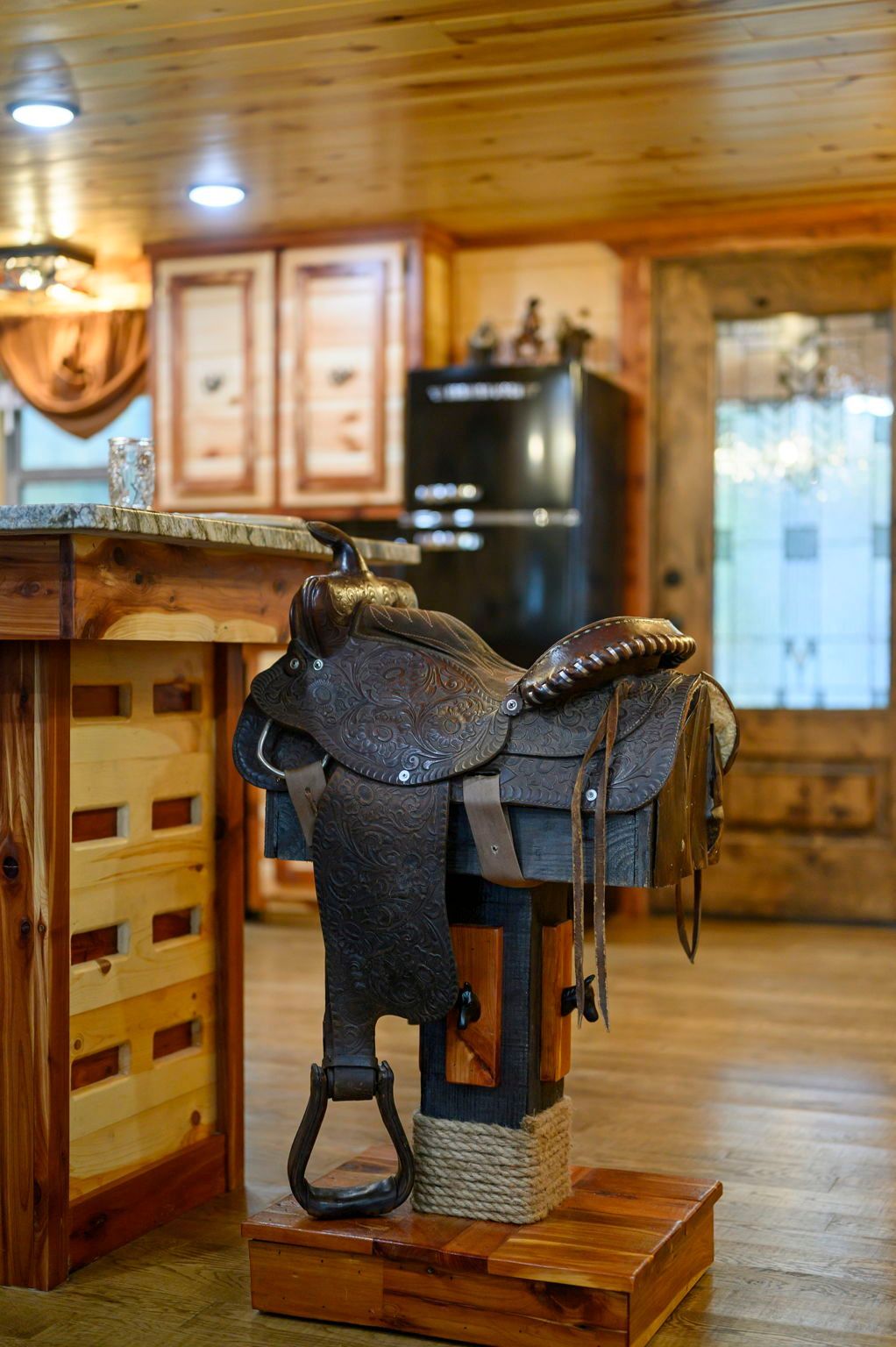 A dark brown leather western saddle rests on a wooden display stand in a rustic, wood-paneled kitchen.