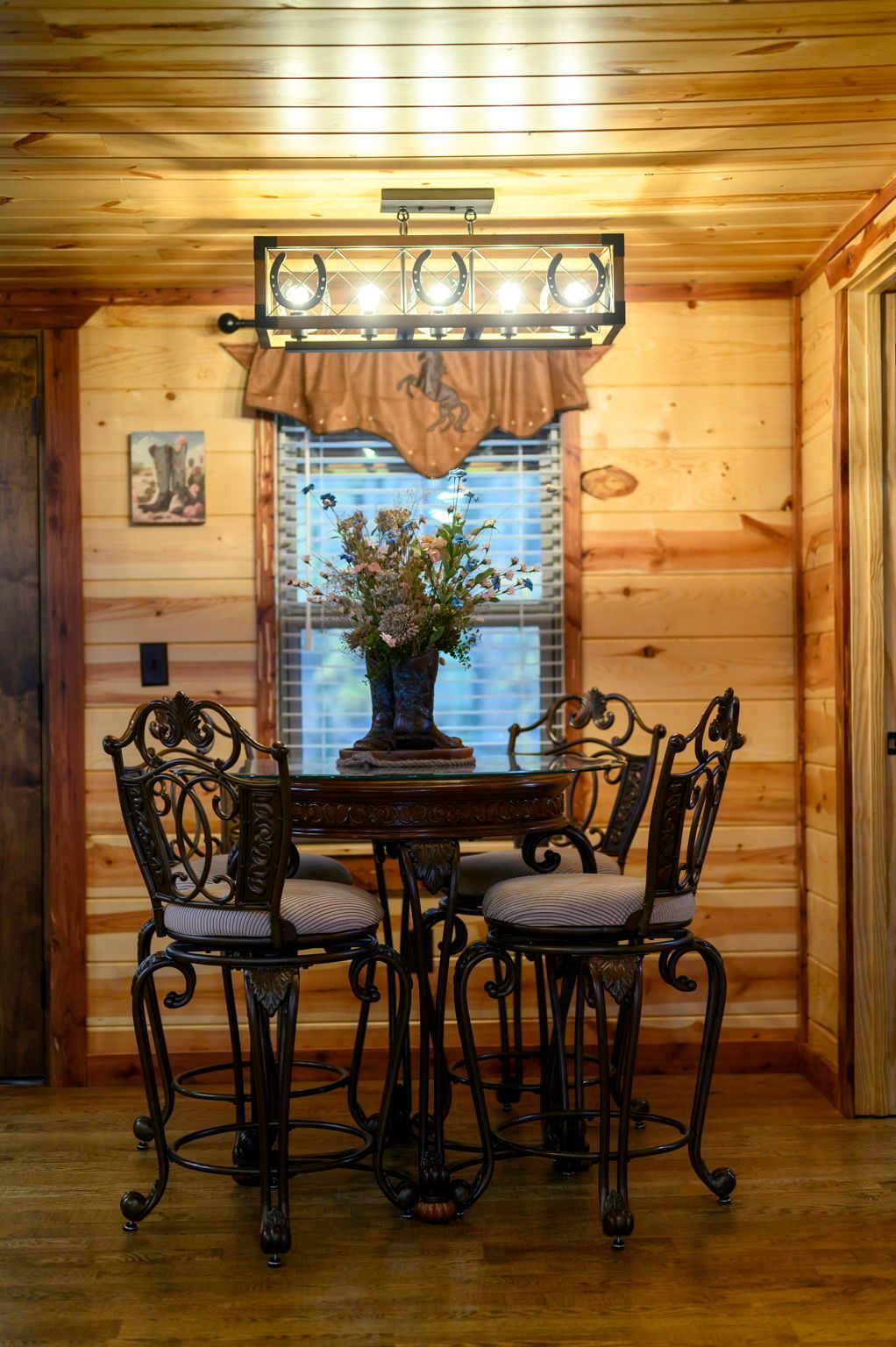 A dining area with a round glass table, four ornate metal chairs, and a rustic horseshoe-themed chandelier in a log room.