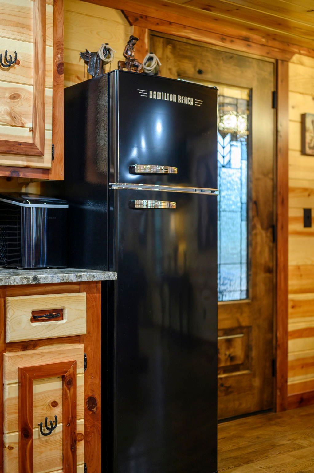 A black retro-style refrigerator stands next to wood cabinets and a rustic wooden door in a room with wood-paneled walls.