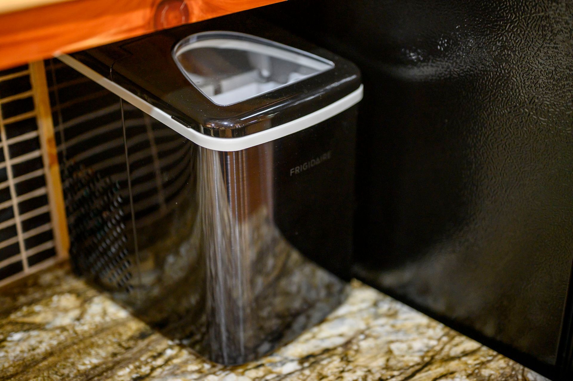 A black portable ice maker sits on a granite countertop under a wooden cabinet, next to a black appliance.