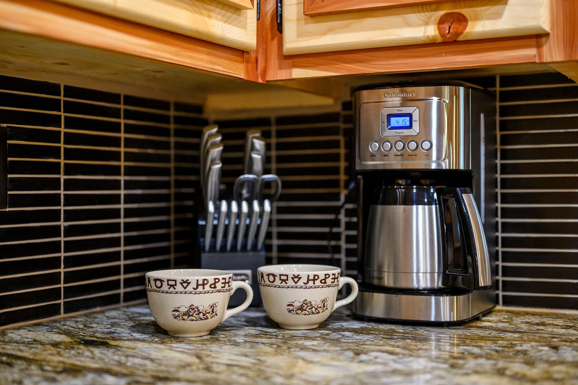 A stainless steel coffee maker with two patterned mugs sits on a granite countertop in a kitchen with a tiled backsplash.