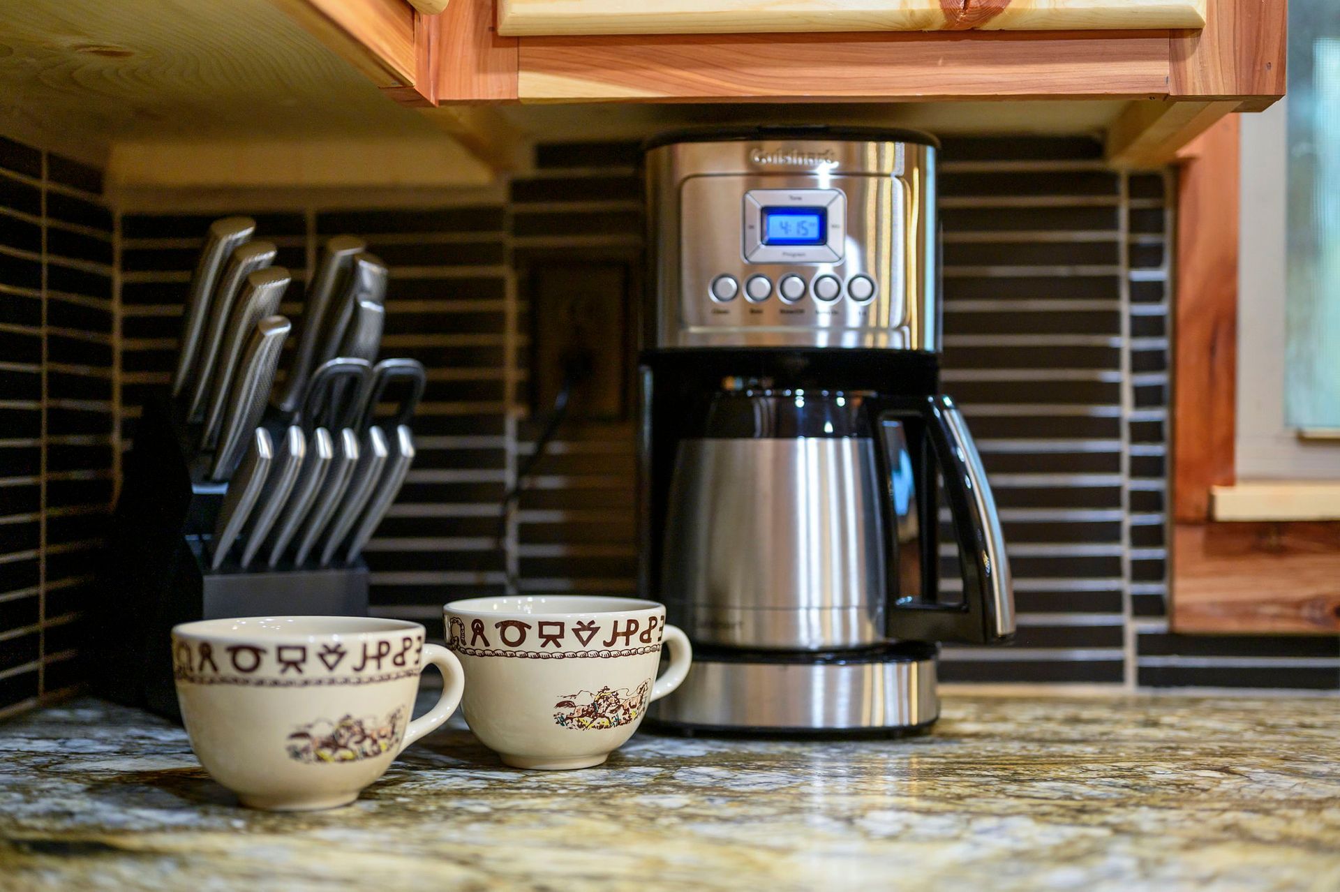 A stainless steel coffee maker sits on a granite countertop next to a knife block and two patterned ceramic coffee mugs.