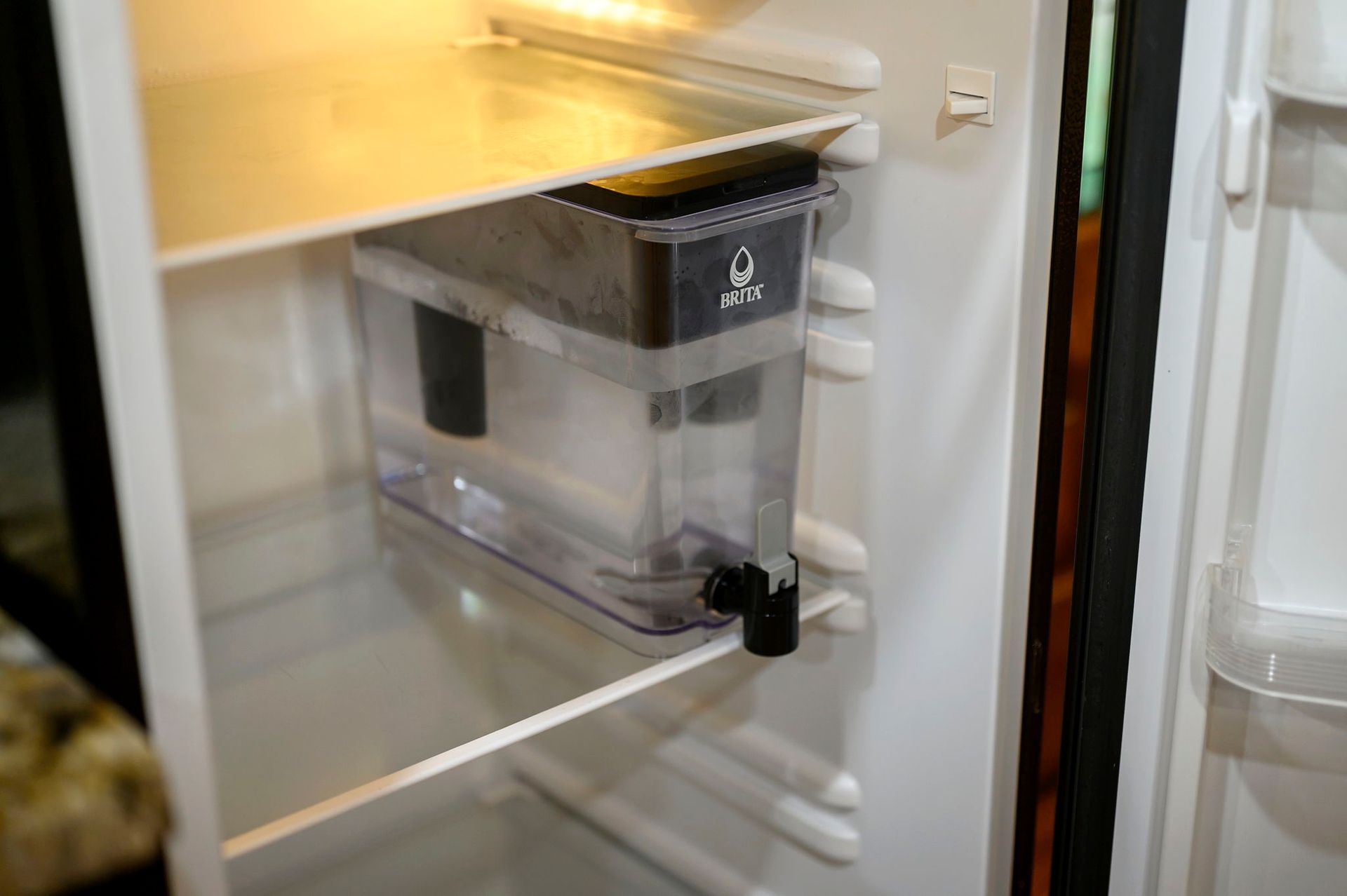 A clear plastic water filtration dispenser with a black lid and spigot sits on a refrigerator shelf.