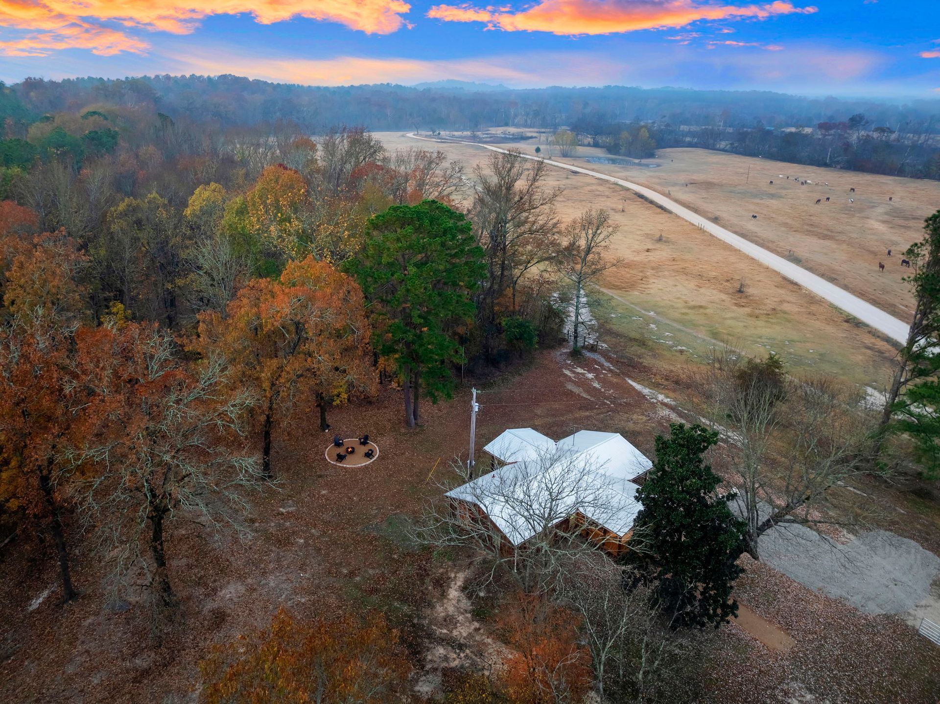 An aerial view of a cabin nestled in a forested area next to a large open field under a colorful sunset sky.