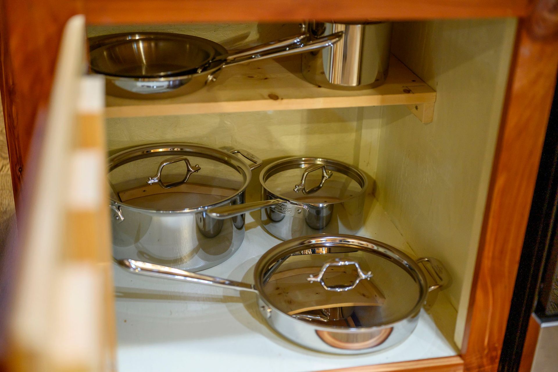 Stainless steel pots and pans with lids organized inside a wooden kitchen cabinet.